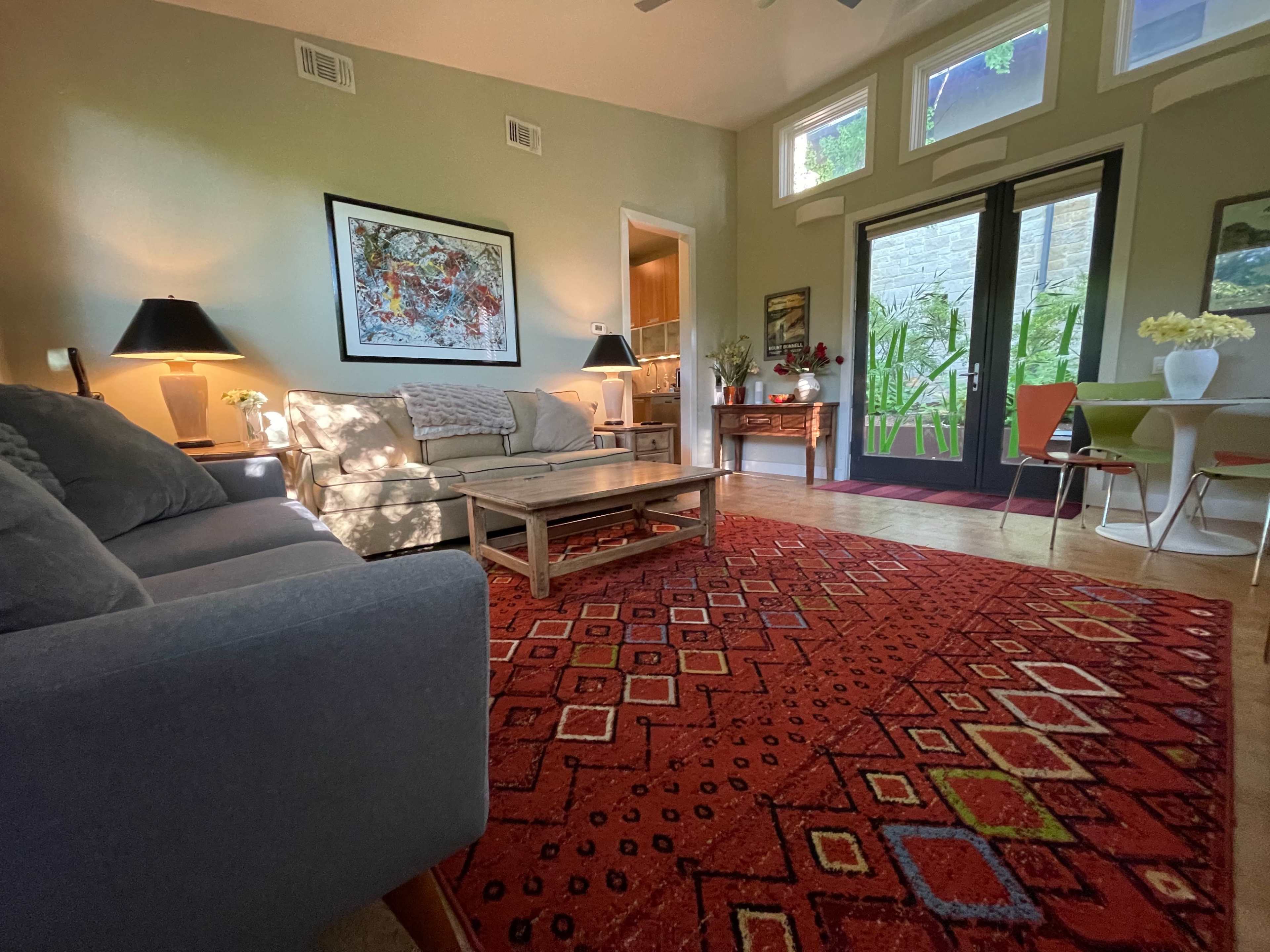 A cozy living room features a gray sofa, a wooden coffee table on a patterned red rug, and large windows overlooking a patio area.
