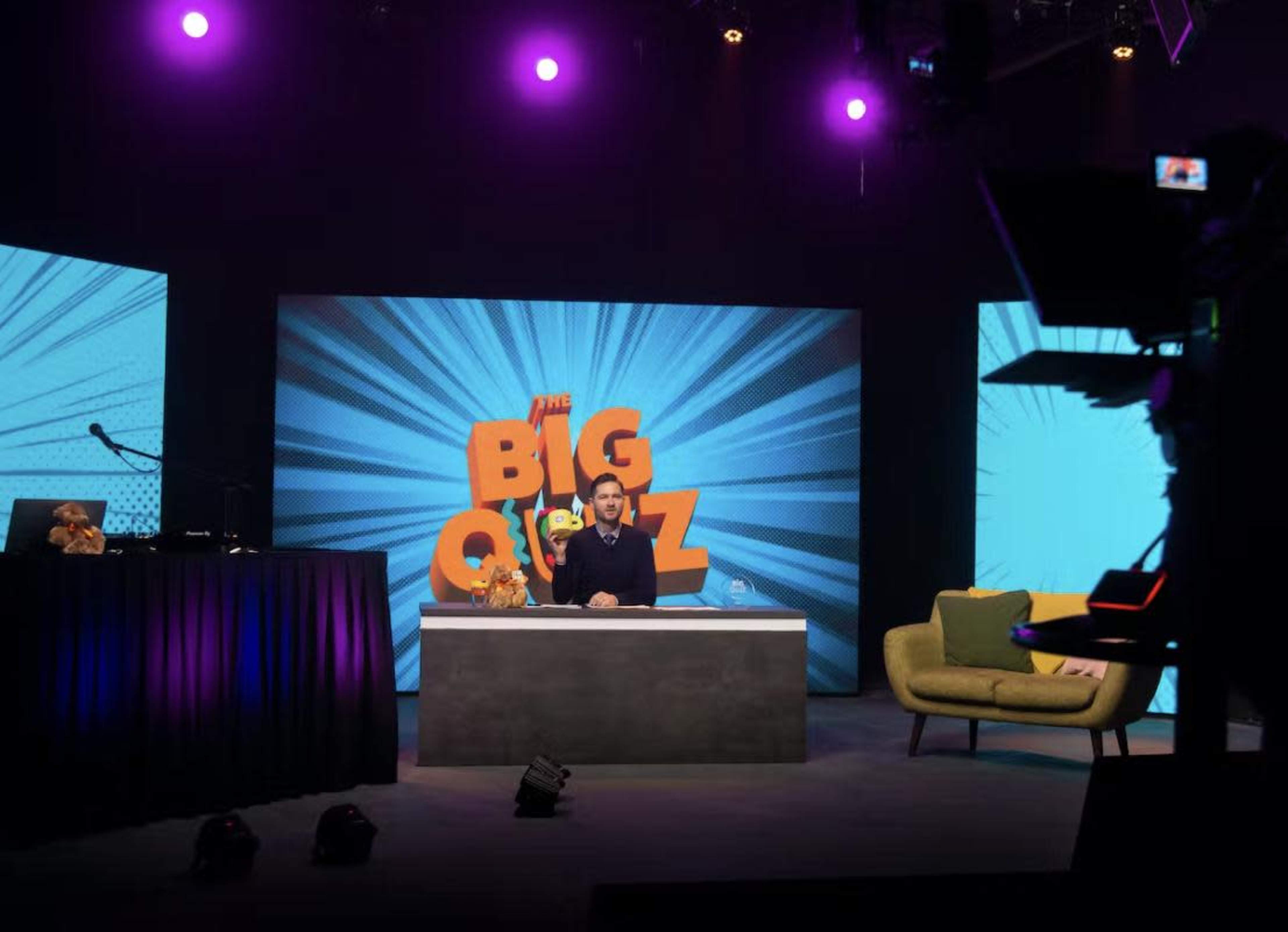 A quiz show setup features a host sitting at a desk with colorful screens behind him and a couch in the foreground.