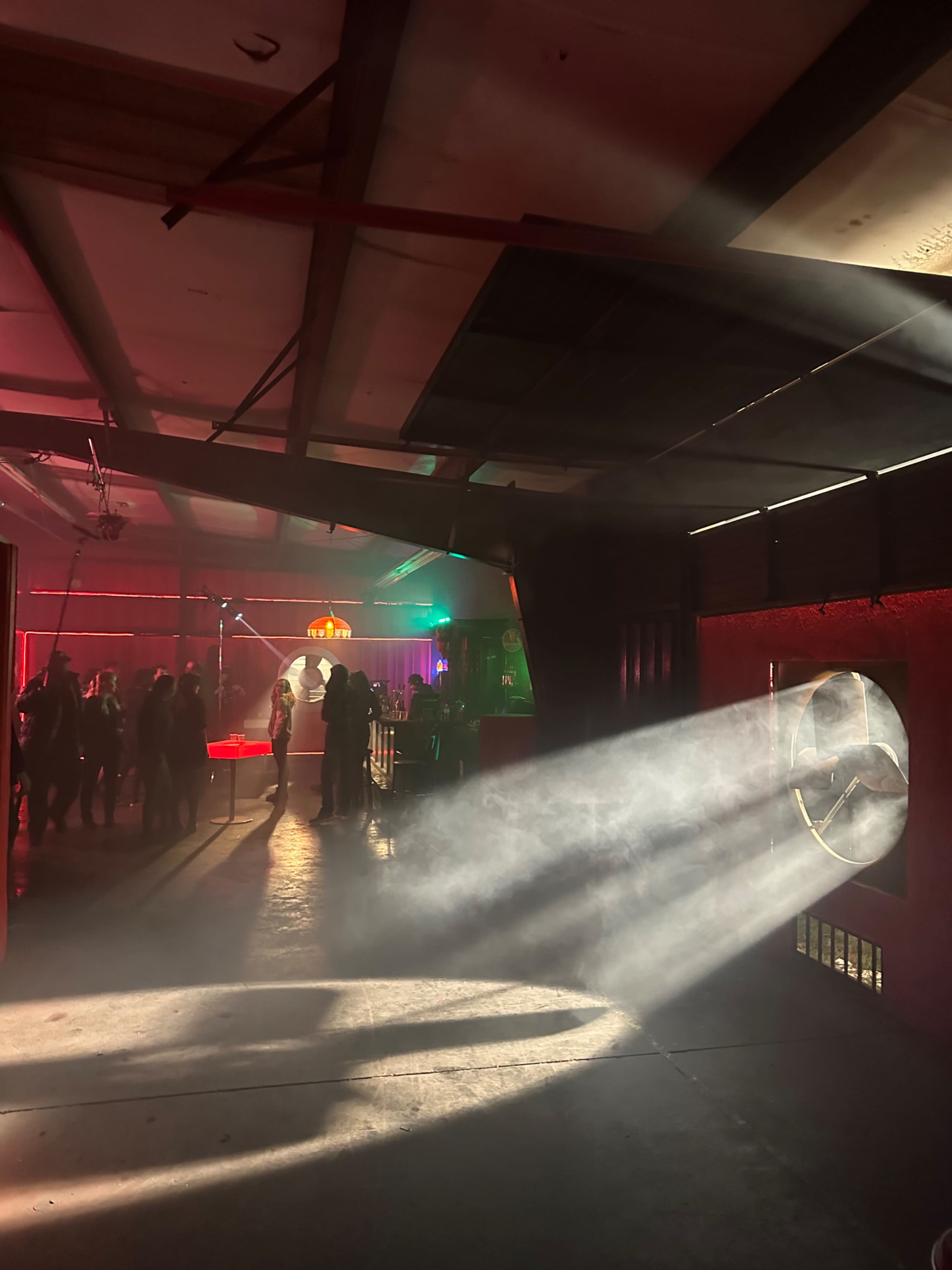 A dimly lit interior with smoke and beams of light streaming through a circular opening, illuminating a gathering of people in the background.