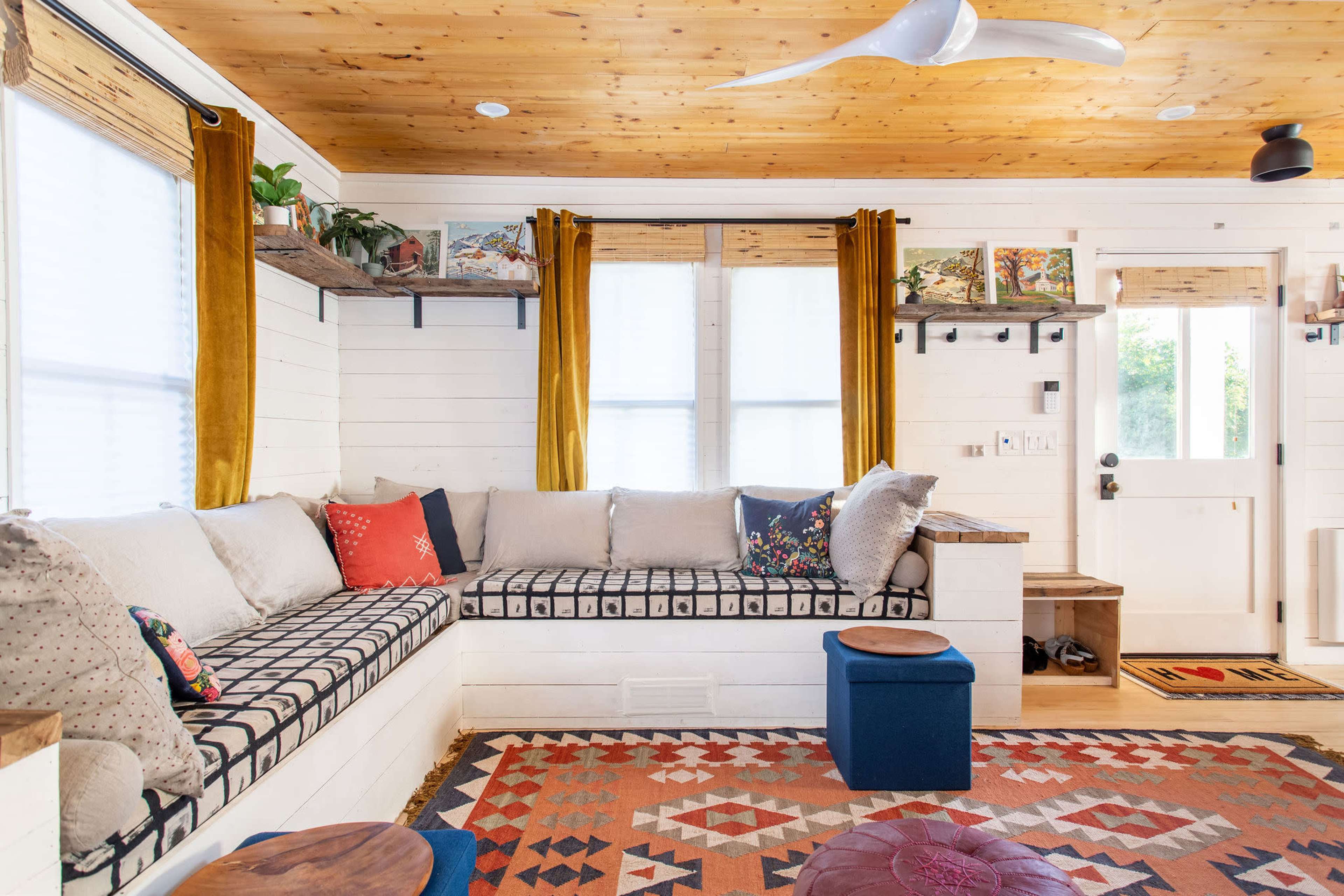 A cozy living room with a built-in seating area, wooden ceiling, decorative rugs, and bright curtains.