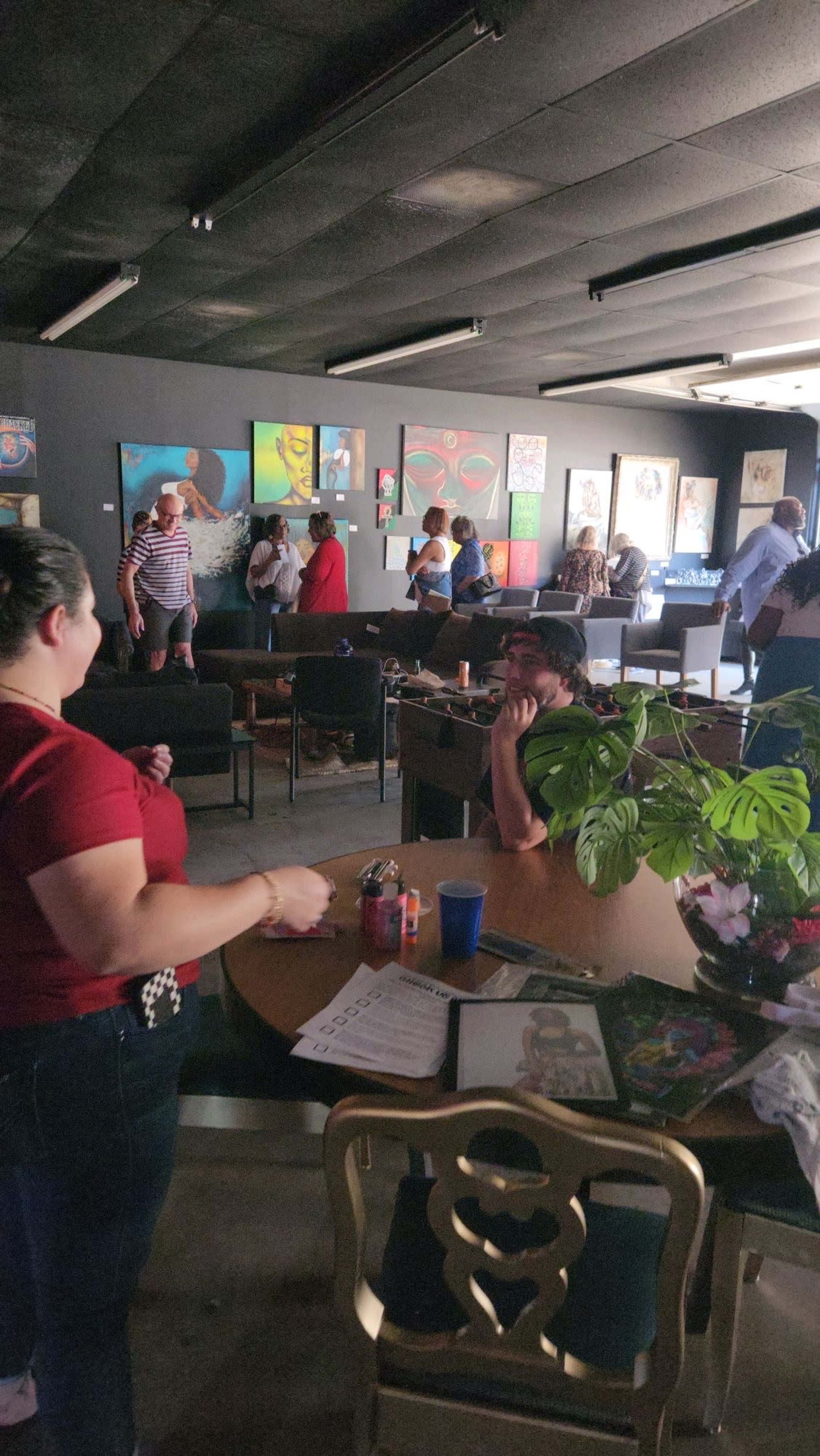 The image shows a lively indoor gathering with people conversing and various artworks displayed on the walls.