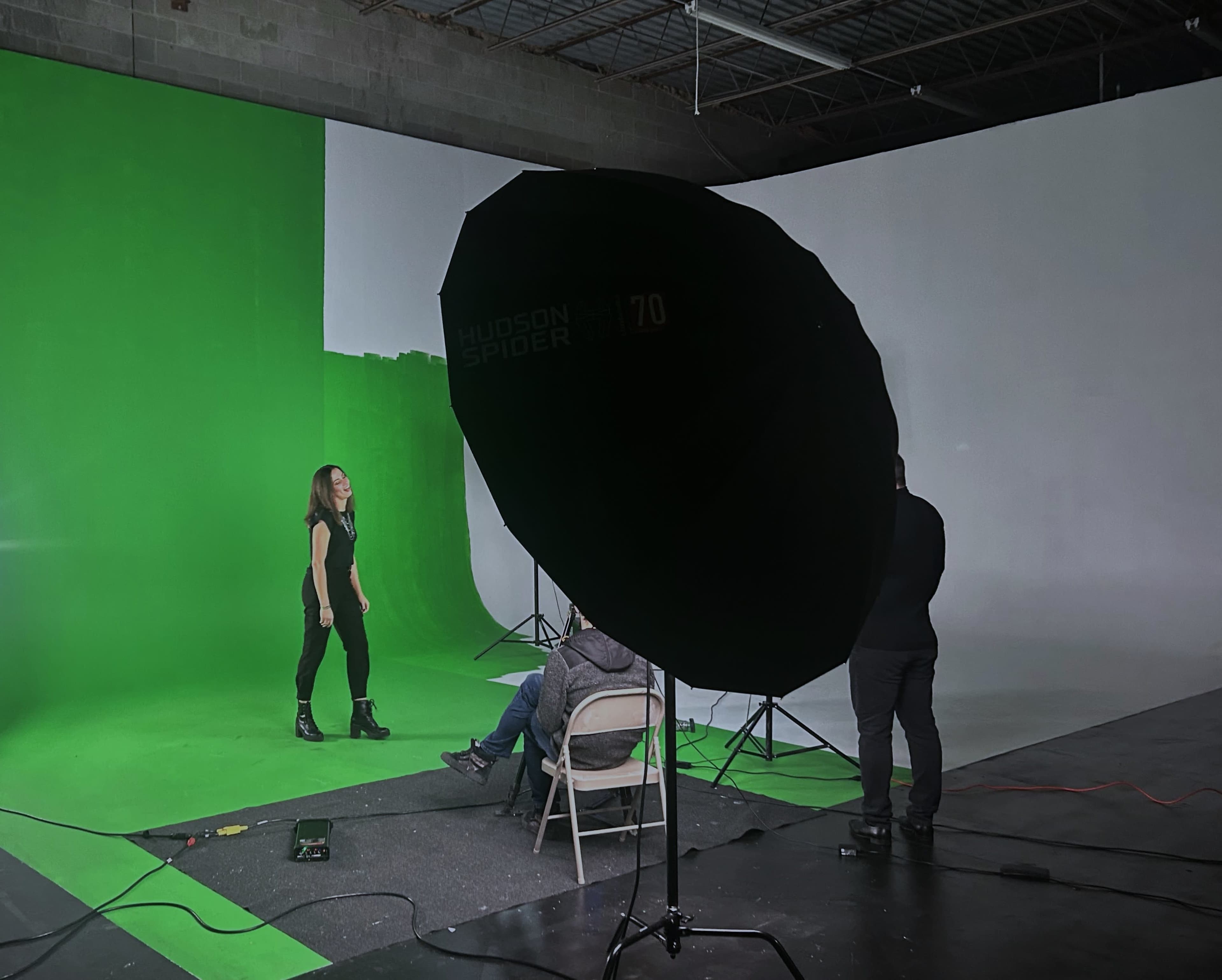 A model stands on a green backdrop while photographers adjust lighting equipment in a studio.