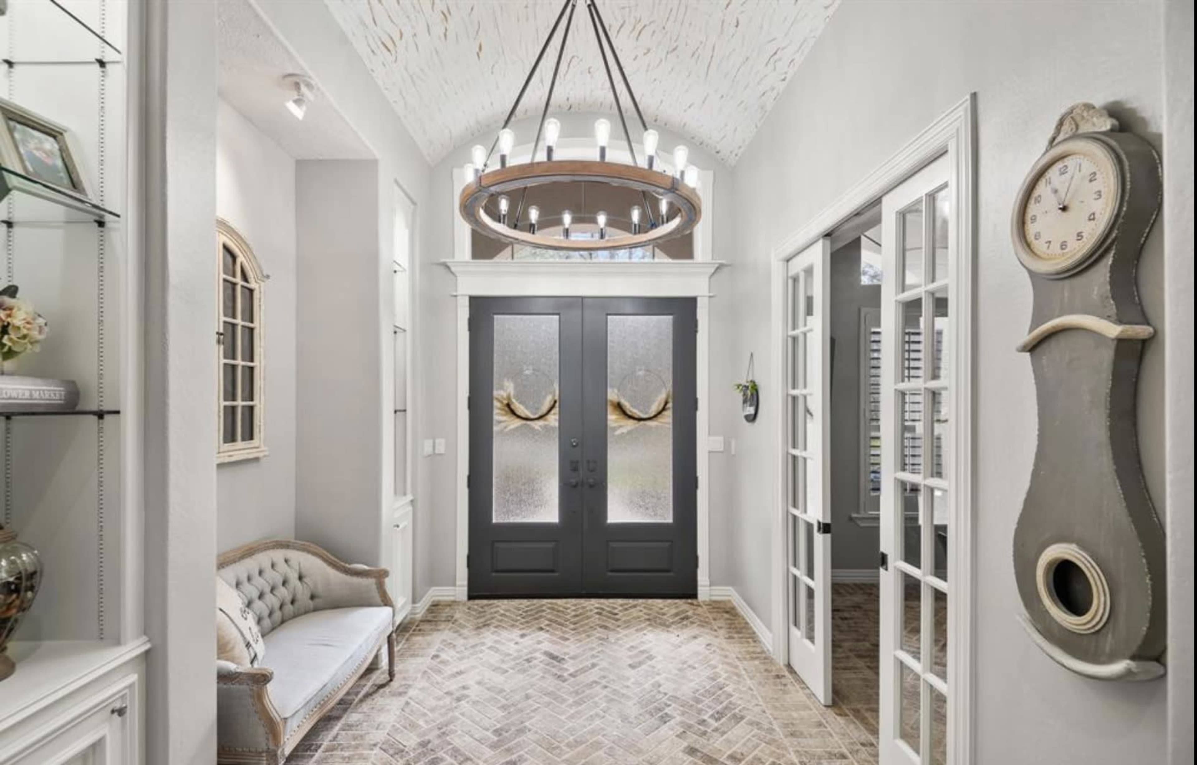 An entrance hall with double doors, a circular chandelier overhead, and a decorative clock on the wall.