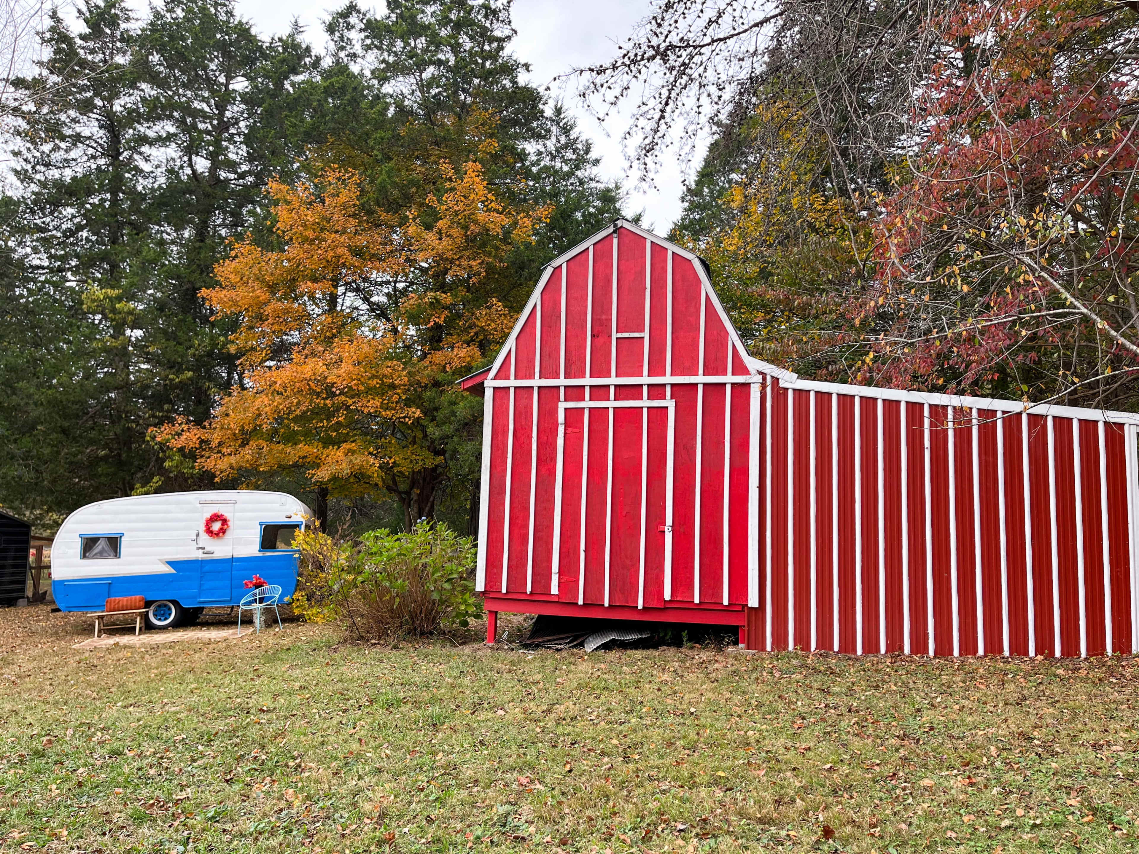 A red and white striped barn stands next to a blue and white vintage trailer, surrounded by autumn foliage.