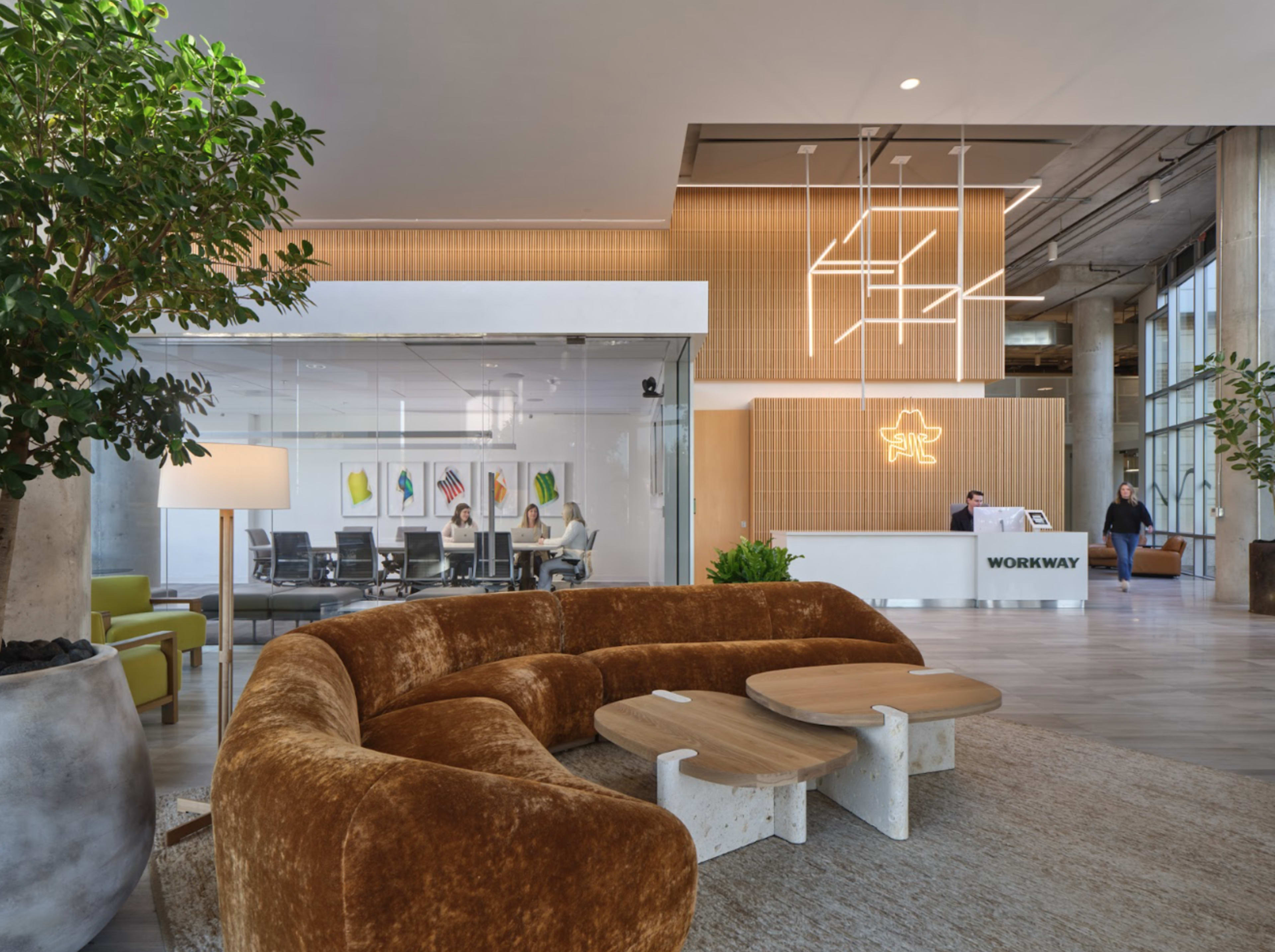 The image shows a modern office reception area featuring a curved brown sofa, a small table set, and a front desk with a neon sign, alongside a meeting room visible through glass walls.