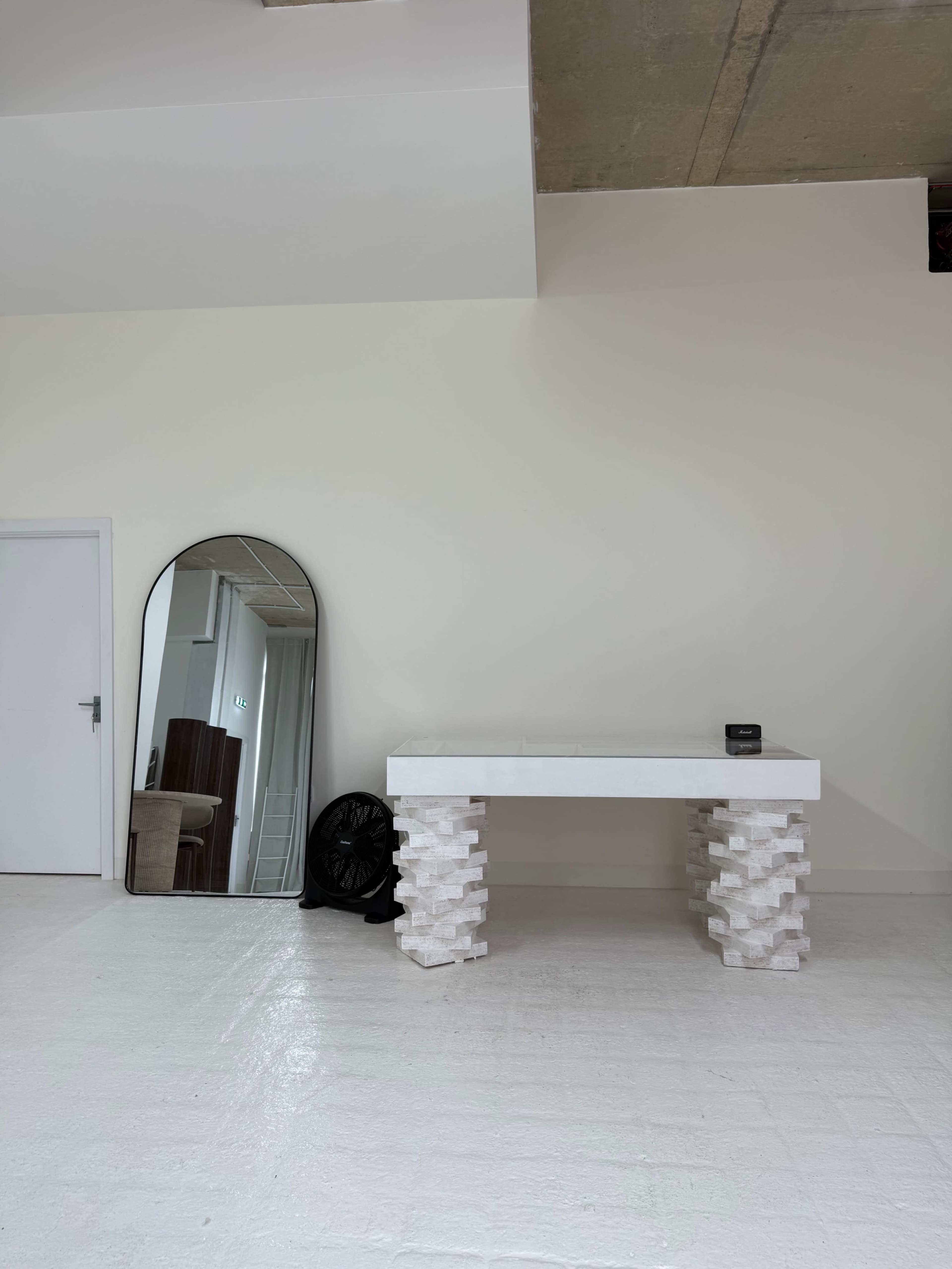 The image shows a minimalist room featuring a white table with textured legs, a large oval mirror against the wall, and a black fan in the corner.