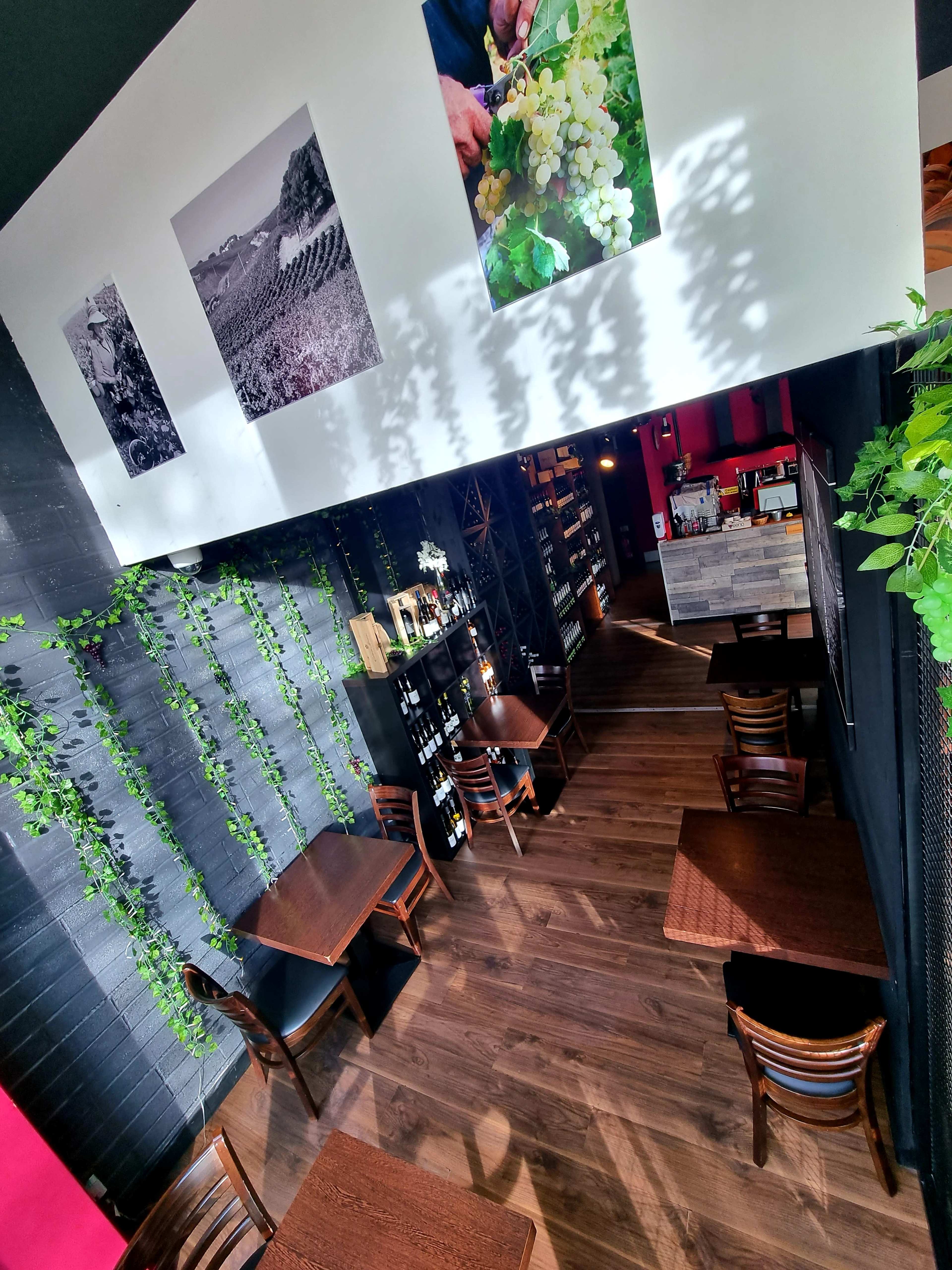 A cozy wine bar with wooden tables, green plants on the walls, and photos of vineyards hanging above.