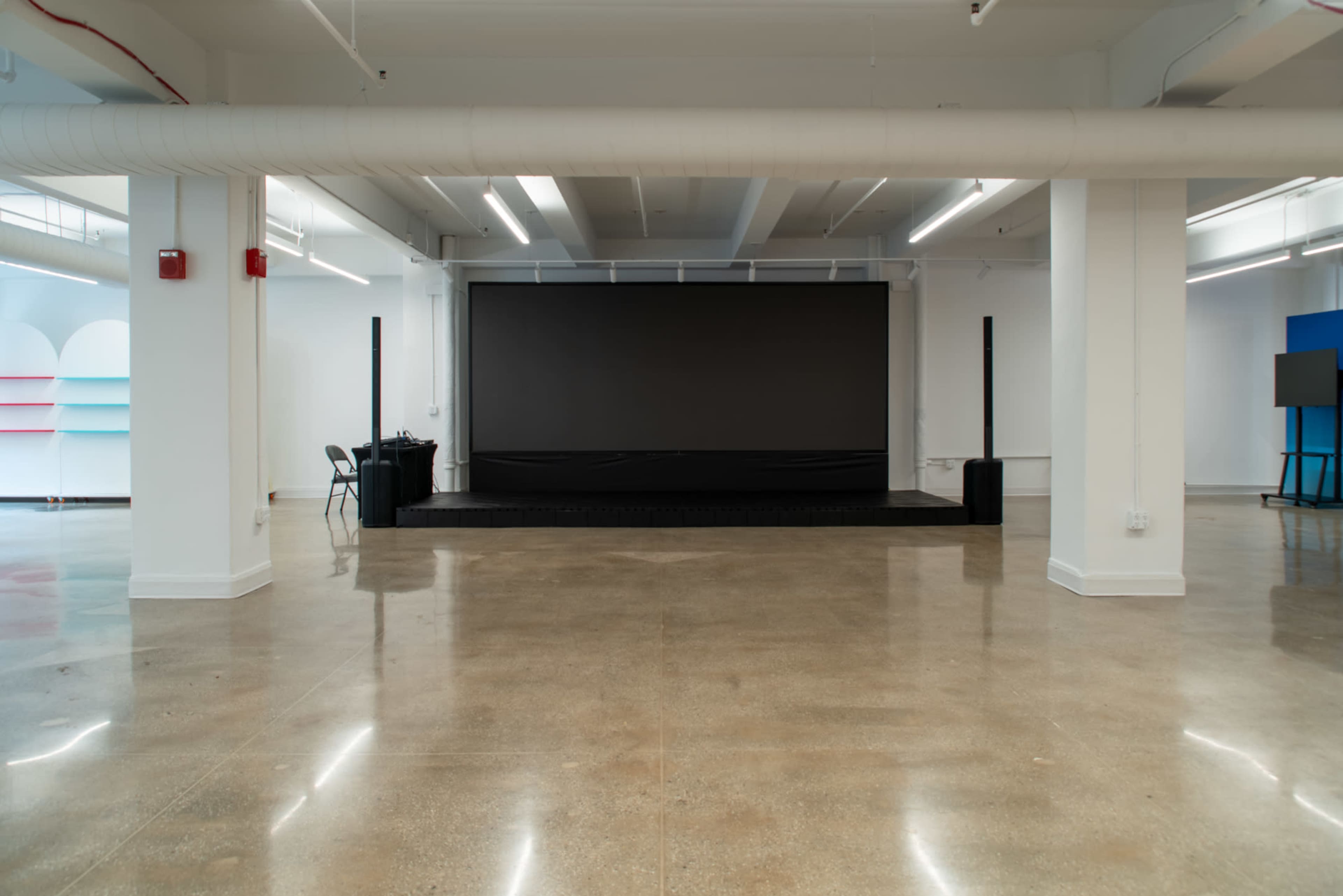 The image shows a spacious, empty room featuring a large black screen at the front, with a polished concrete floor and minimal furniture.