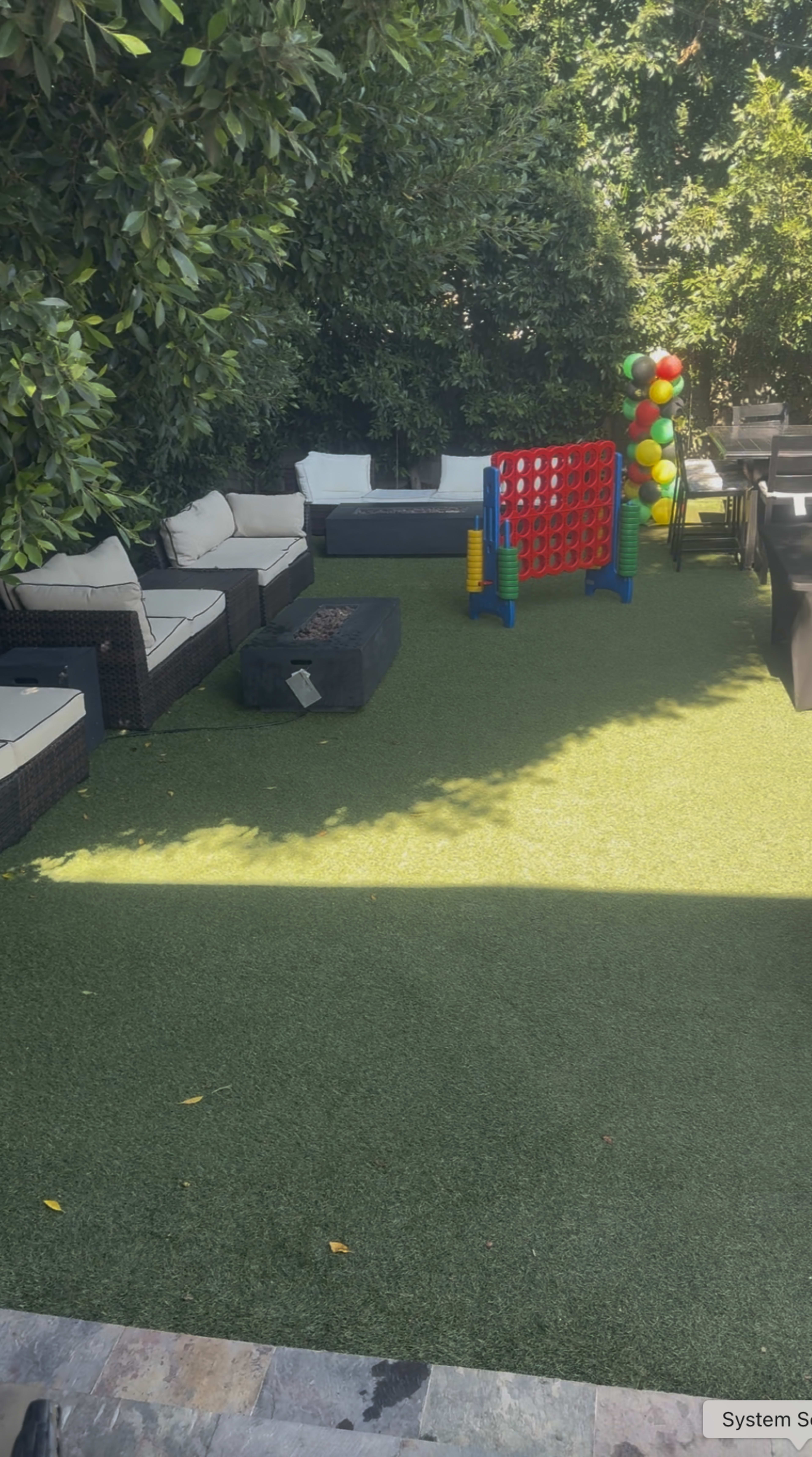 The image shows a backyard garden with seating areas, a colorful game structure, and a barbecue setup on artificial grass.