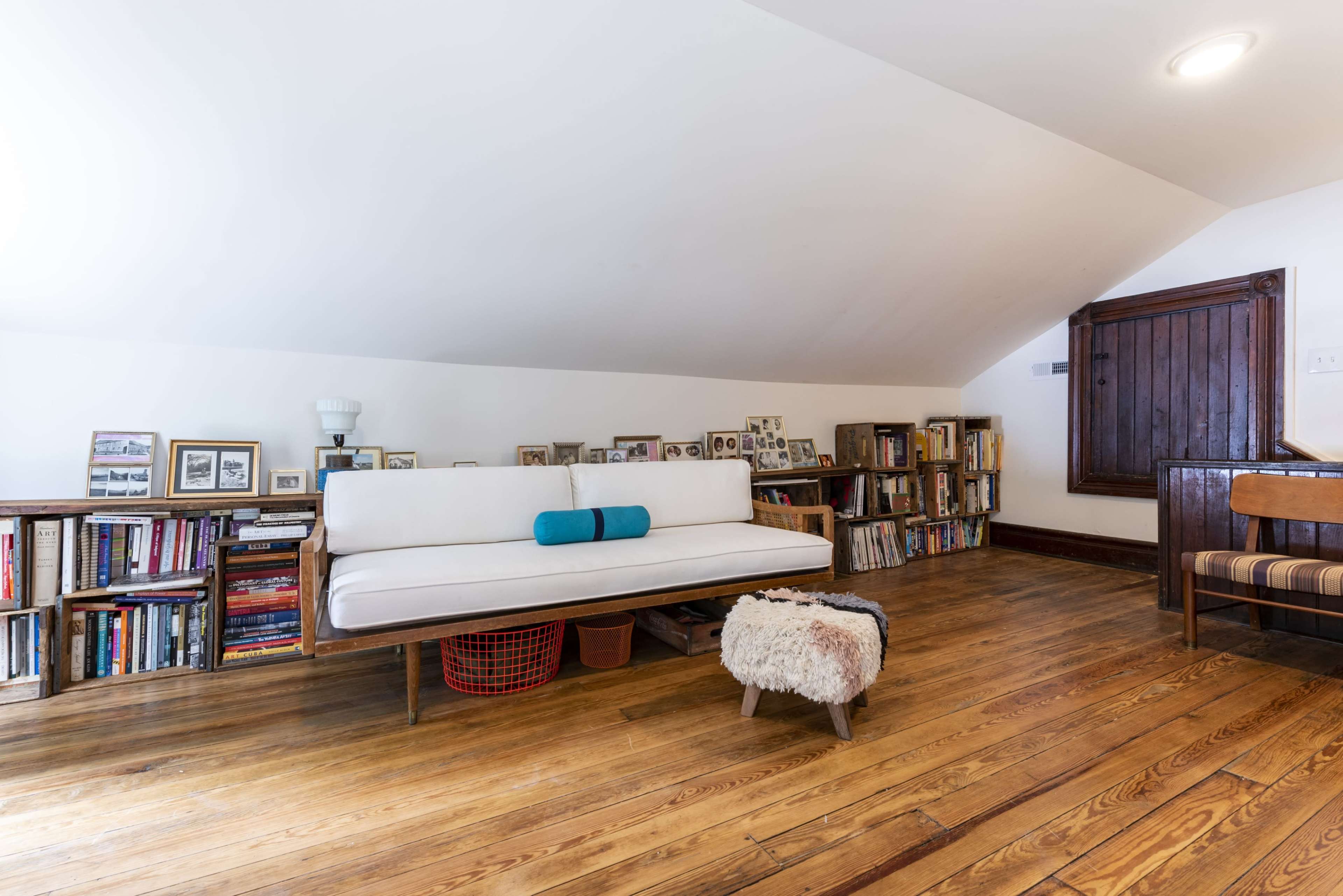 A small attic room features a white sofa, wooden bookshelves filled with books and framed photographs, and a wooden floor.