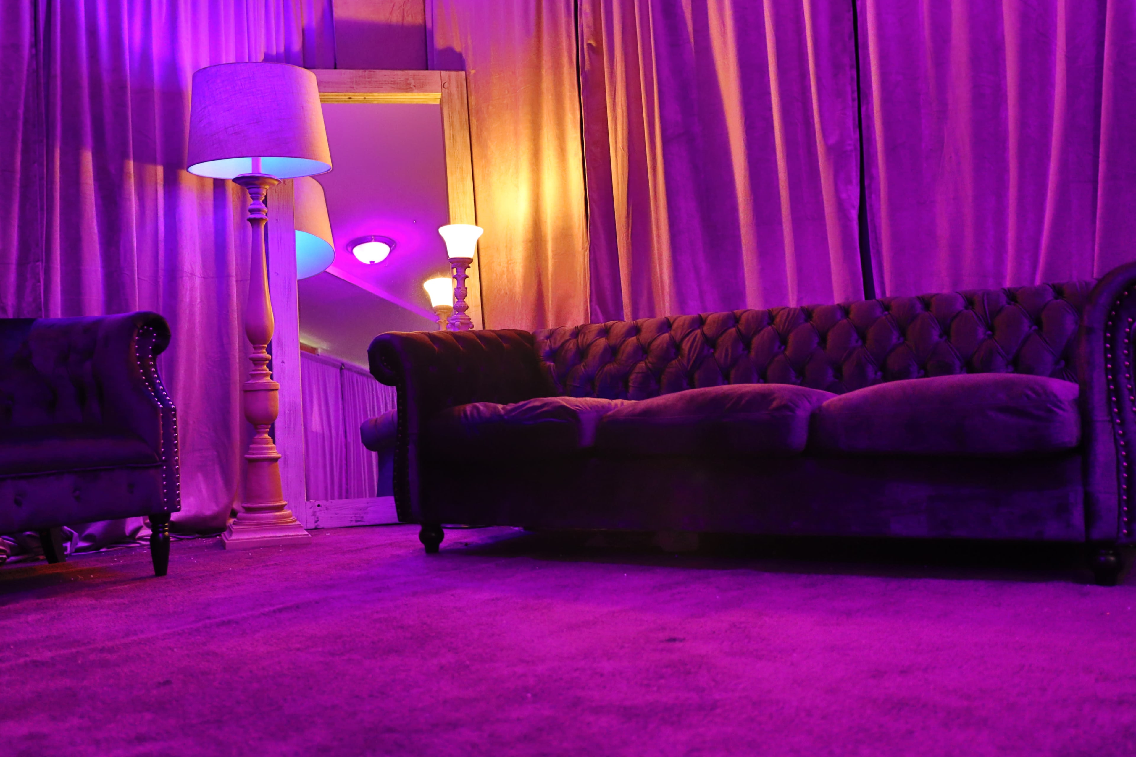 A velvet sofa and armchair are positioned in a room with purple curtains and ambient lighting from lamps.