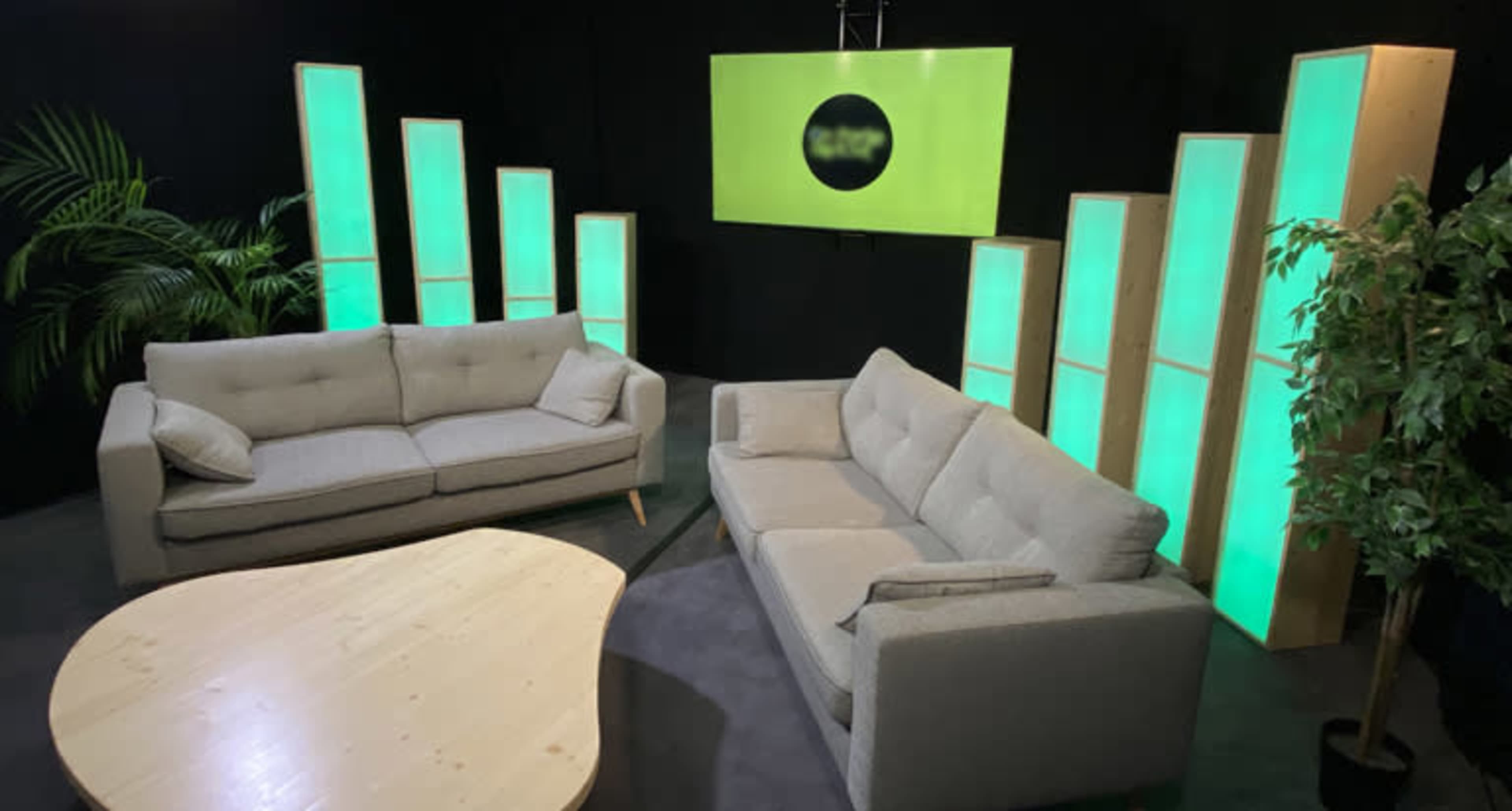 A minimalist lounge area with two gray sofas arranged around a light-colored wooden coffee table, set against a backdrop of illuminated green panels and a screen.