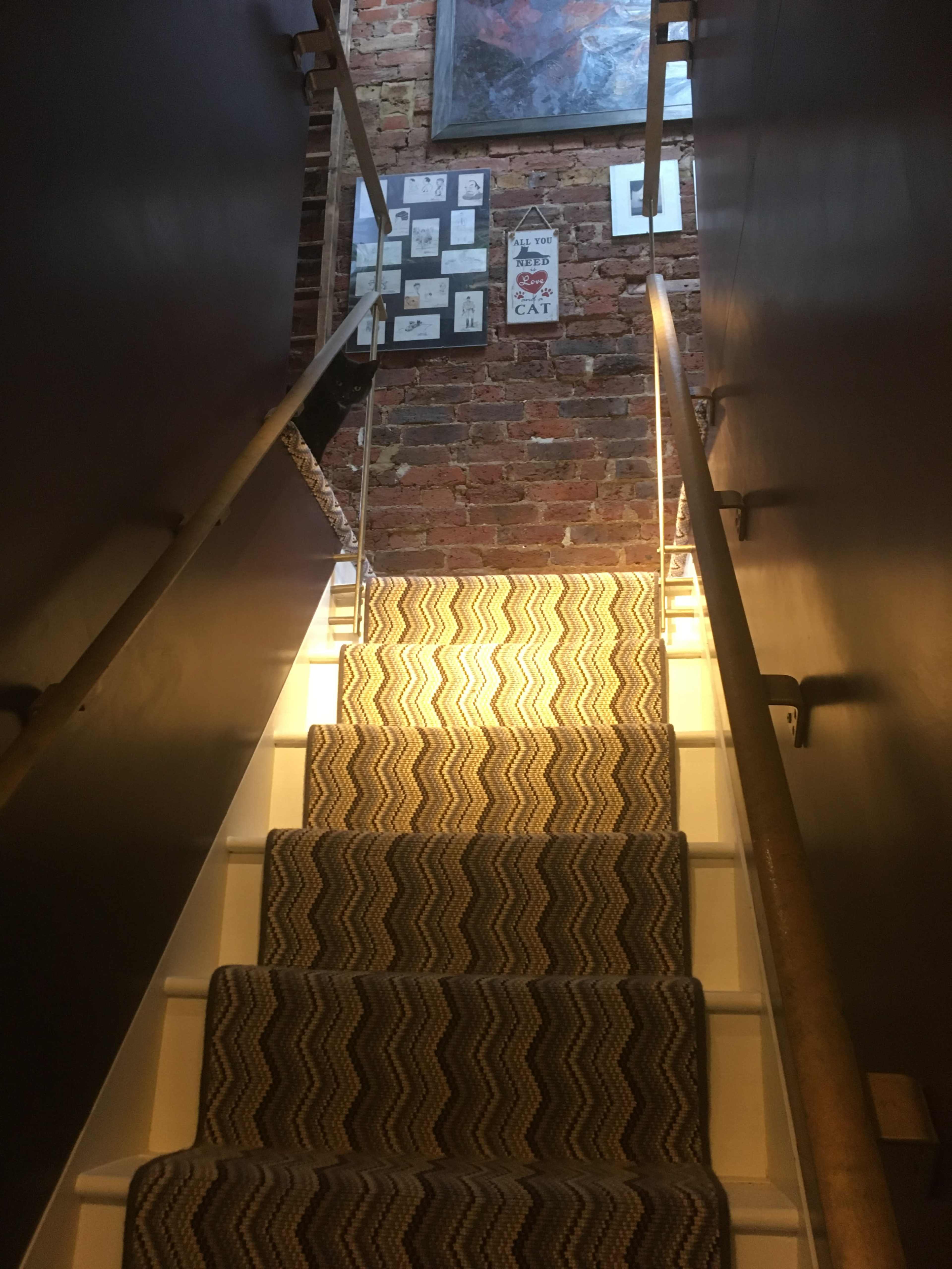 A staircase with a patterned runner leads upward toward a brick wall adorned with framed pictures.