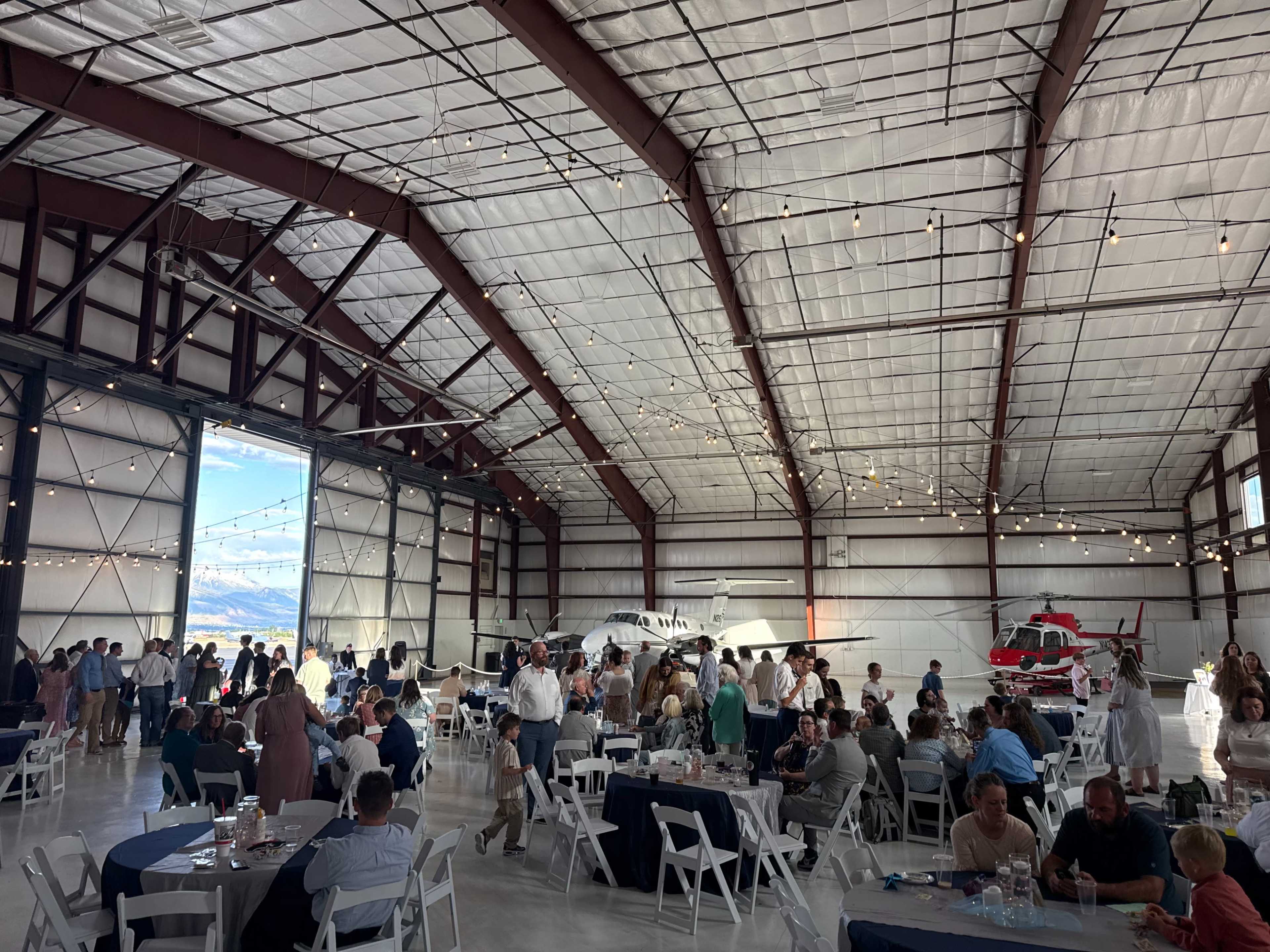 The image shows a large hangar filled with people dining and socializing, with airplanes visible in the background and lights strung across the ceiling.