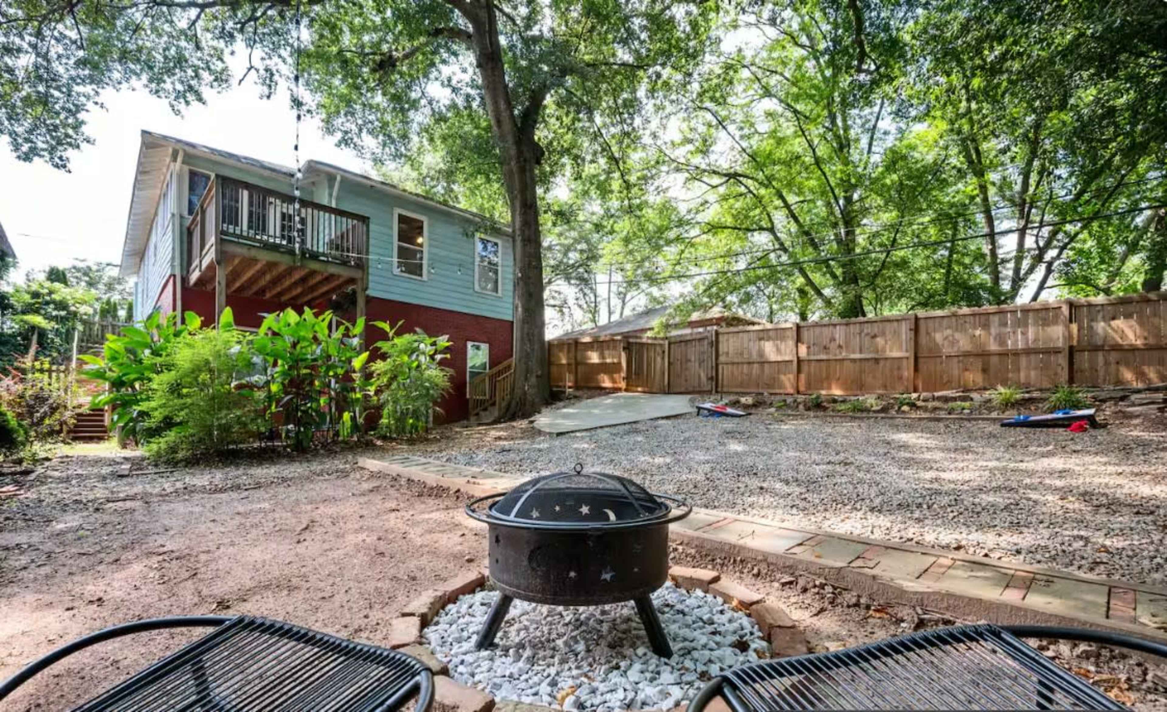 A backyard features a fire pit surrounded by gravel, with a two-story house and a fenced area filled with trees and vegetation in the background.