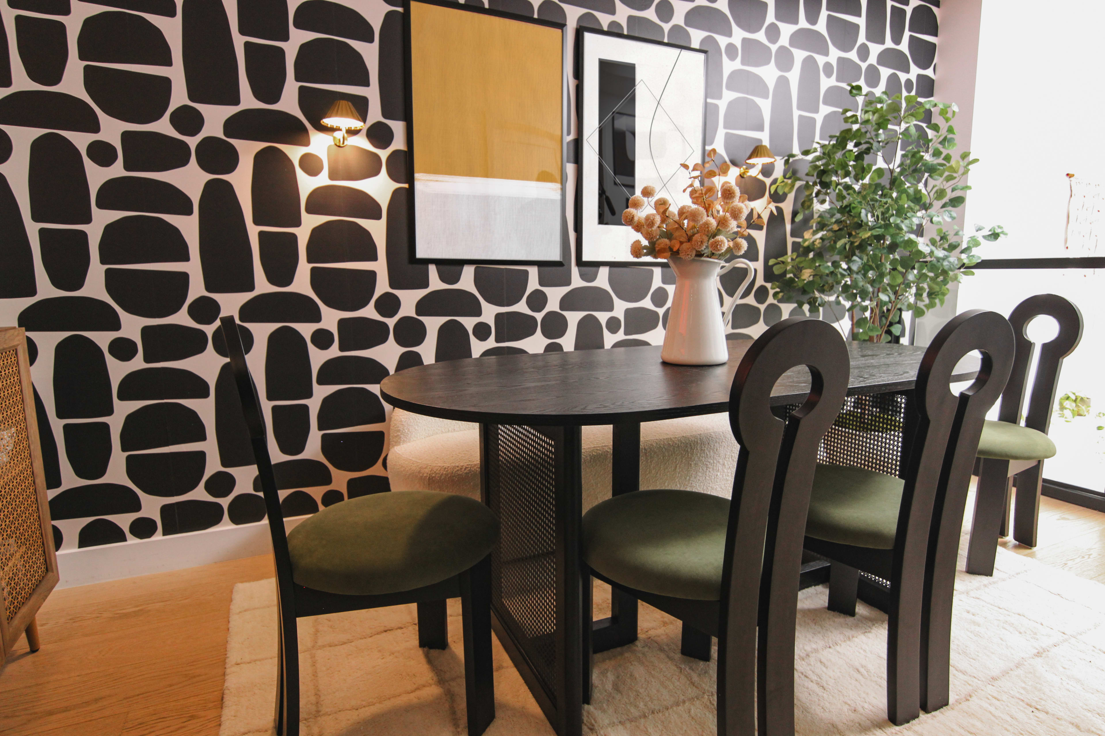 A dining area features a dark wooden table surrounded by four green upholstered chairs, set against a patterned black and white wall and illuminated by wall sconces.