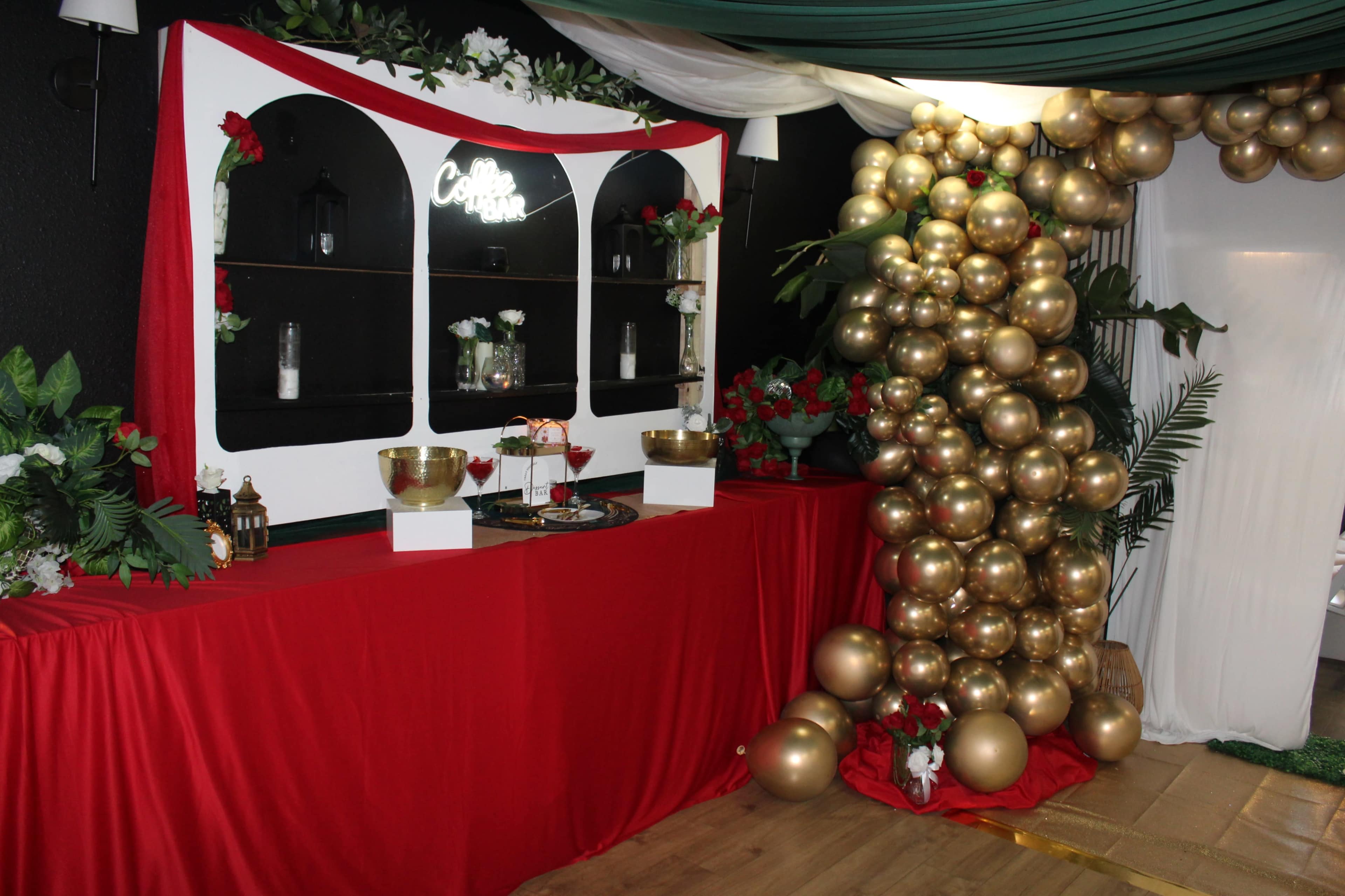The image shows a decorated event space featuring a red table with a backdrop of shelves adorned with plants and lanterns, alongside a large display of gold balloons.