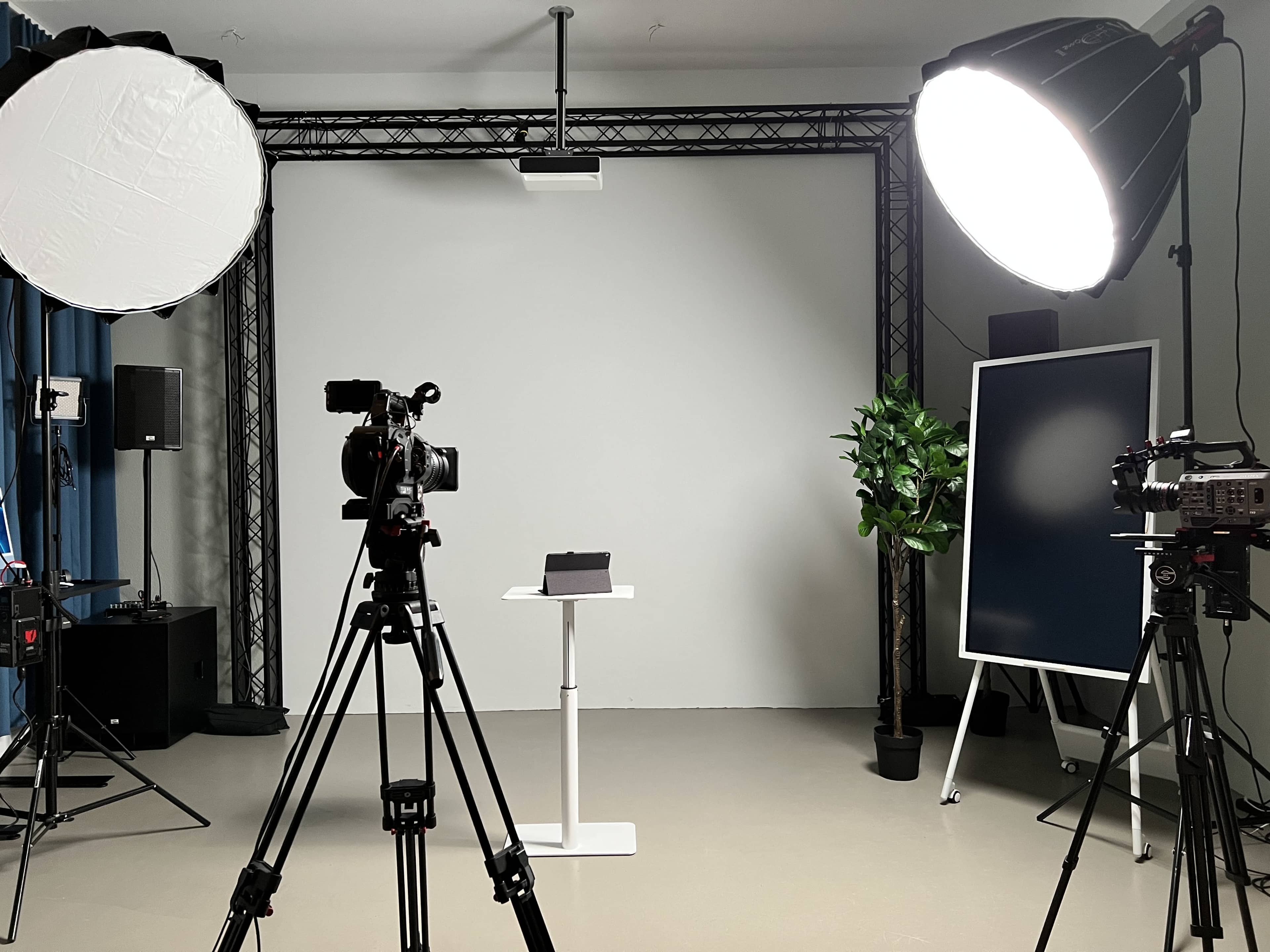 A photography studio setup features two cameras on tripods, a white backdrop, lighting equipment, and a small table with a plant and a black object on it.