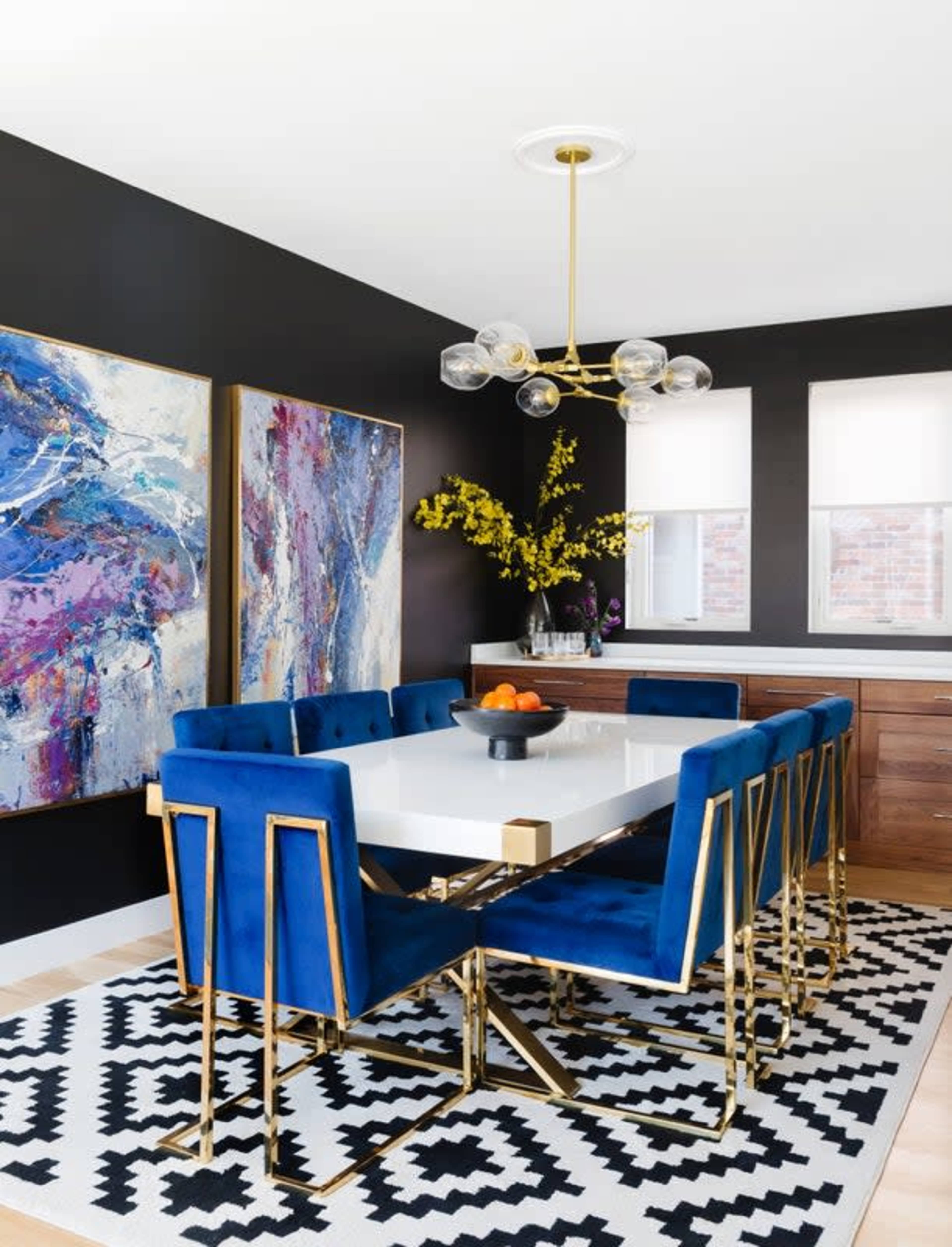 A modern dining room features a white table surrounded by blue velvet chairs, with abstract art on the walls and a decorative centerpiece of fruit.