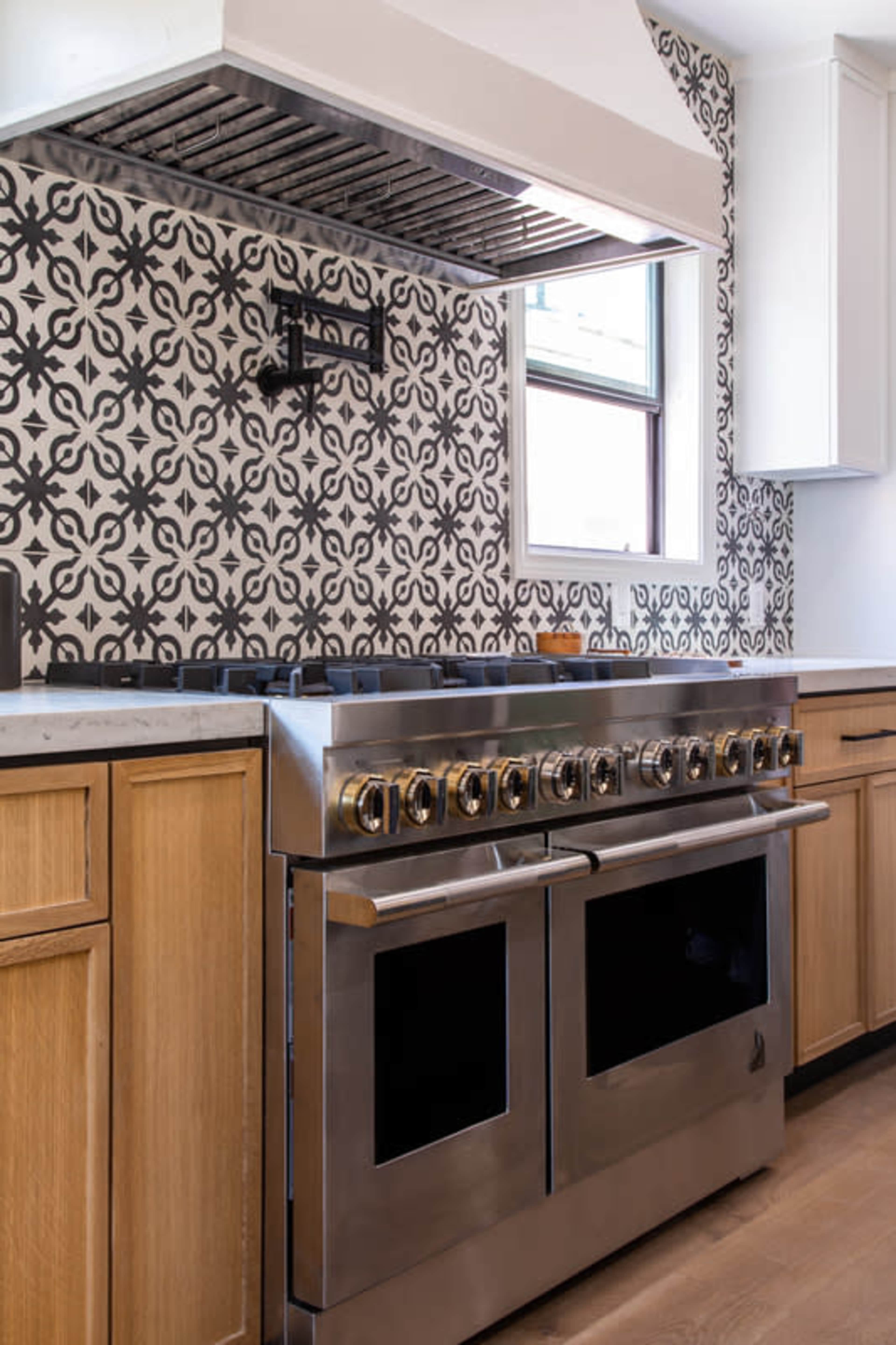A stainless steel gas range with a vent hood is set against a patterned tile backsplash in a kitchen with wooden cabinetry.