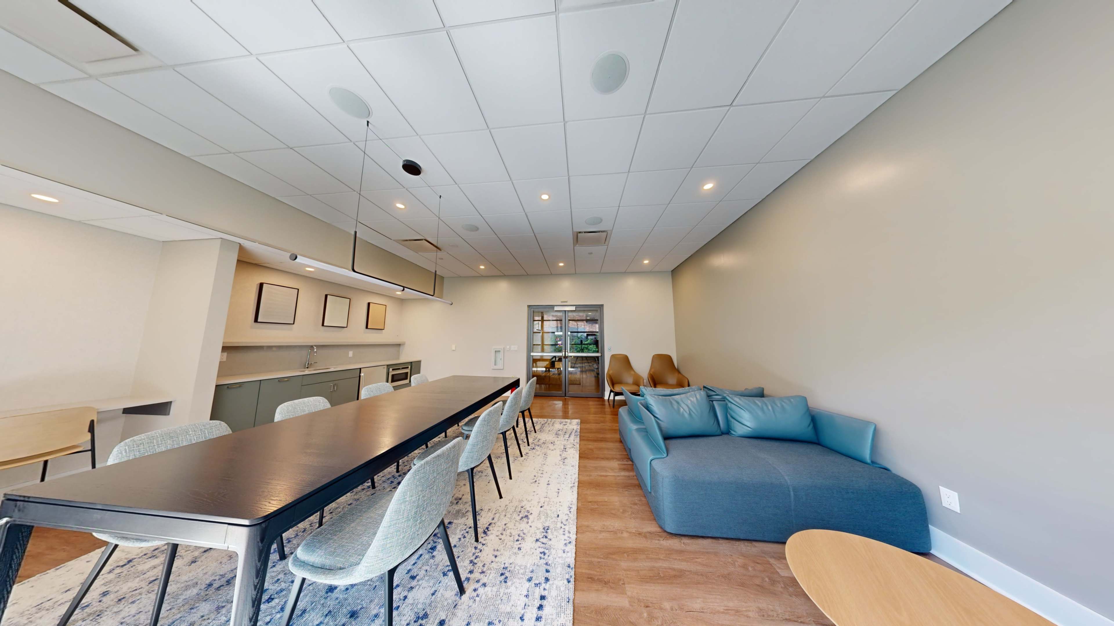 The image shows a modern meeting room featuring a long table with chairs, a cozy seating area with a blue couch, and a kitchen space at the far end.