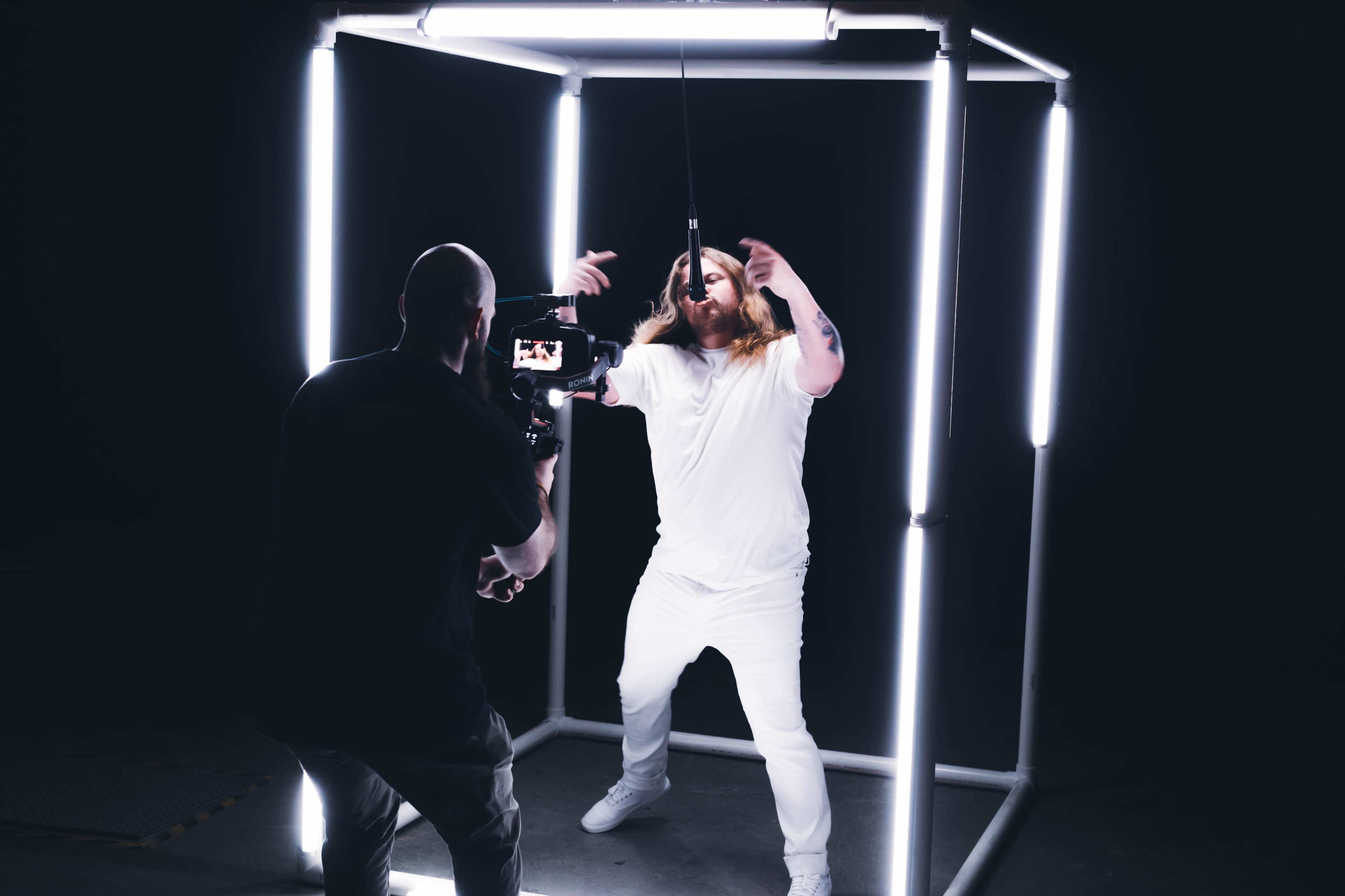 A person in a white outfit performs inside a box of bright lights while being filmed by another individual holding a camera.