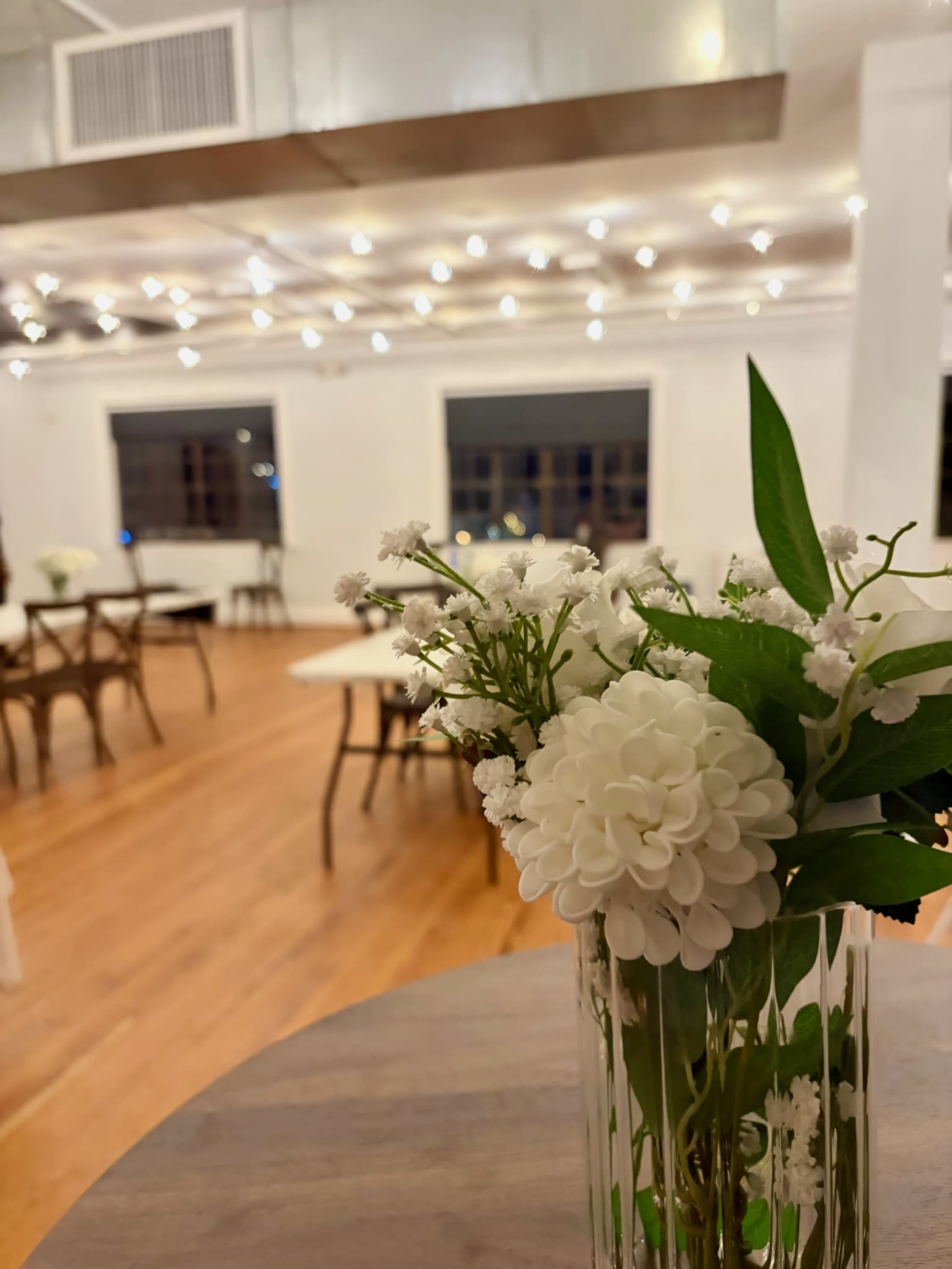 A clear vase of white flowers is placed on a wooden table in an event space with string lights and empty tables in the background.