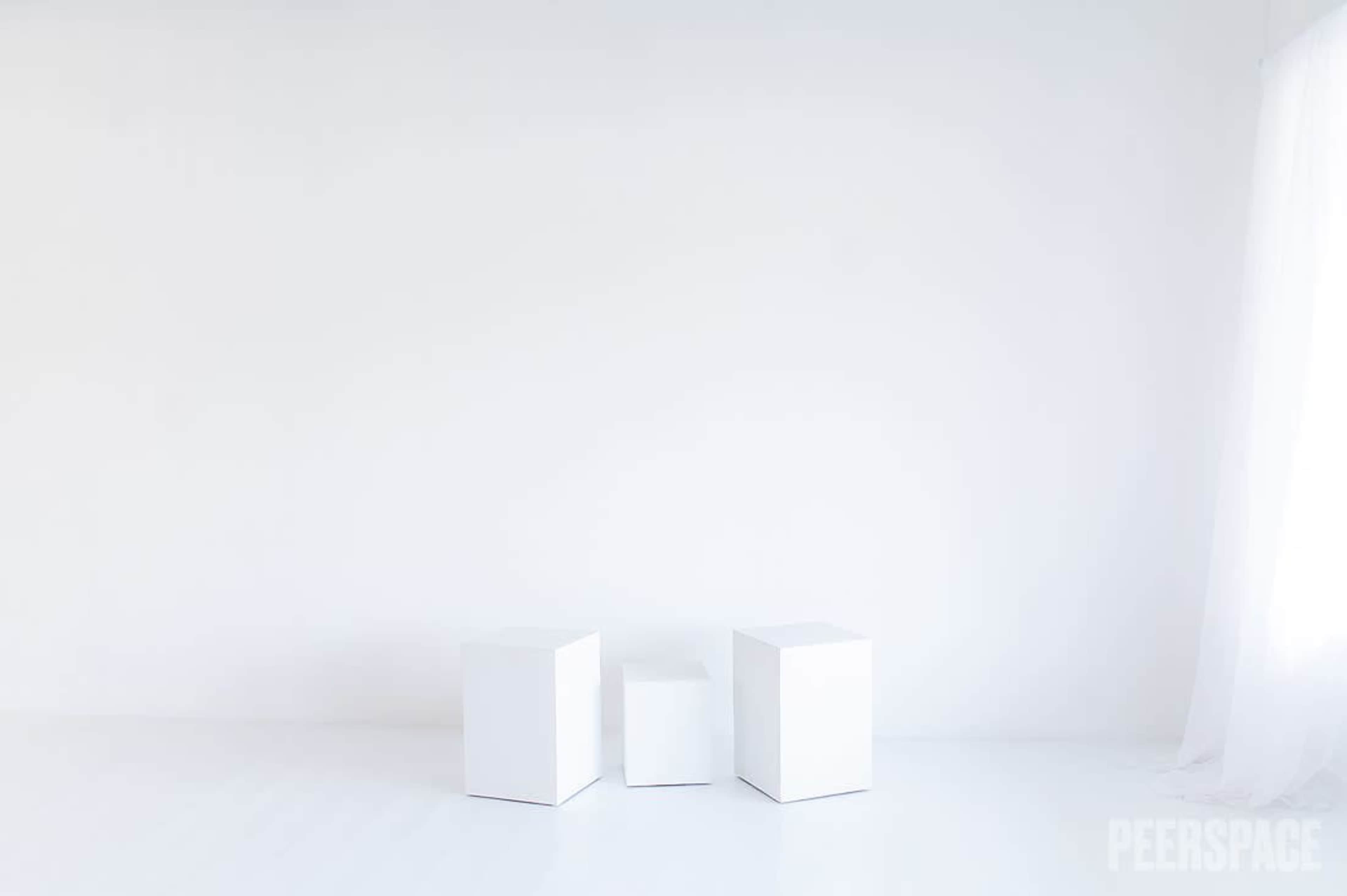 The image shows three white geometric cubes arranged in a minimalist white room with a sheer curtain.