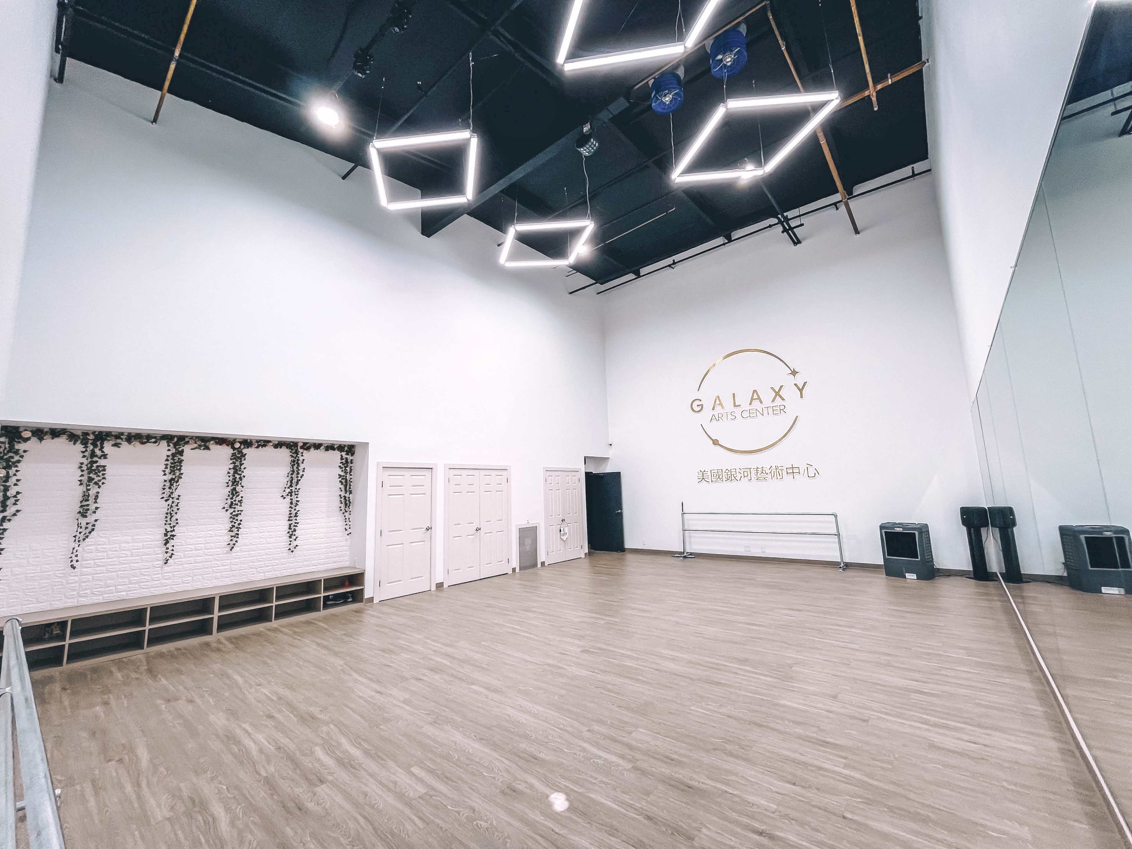 A spacious dance studio with a mirrored wall, wooden flooring, and minimalist decor, featuring modern lighting fixtures.