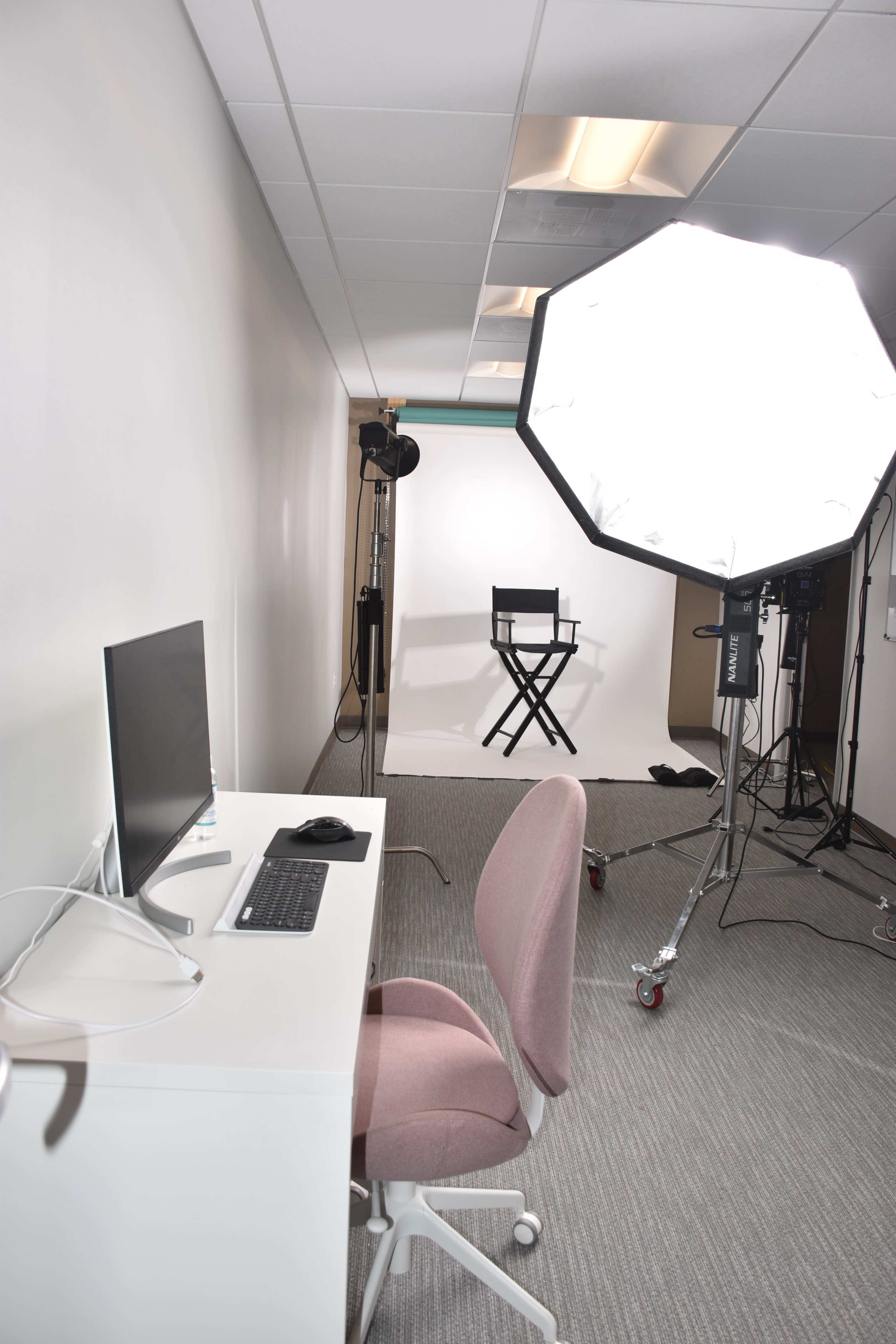 A clean, modern office space with a desk, computer, and a chair facing a brightly lit area with a backdrop and a director's chair.