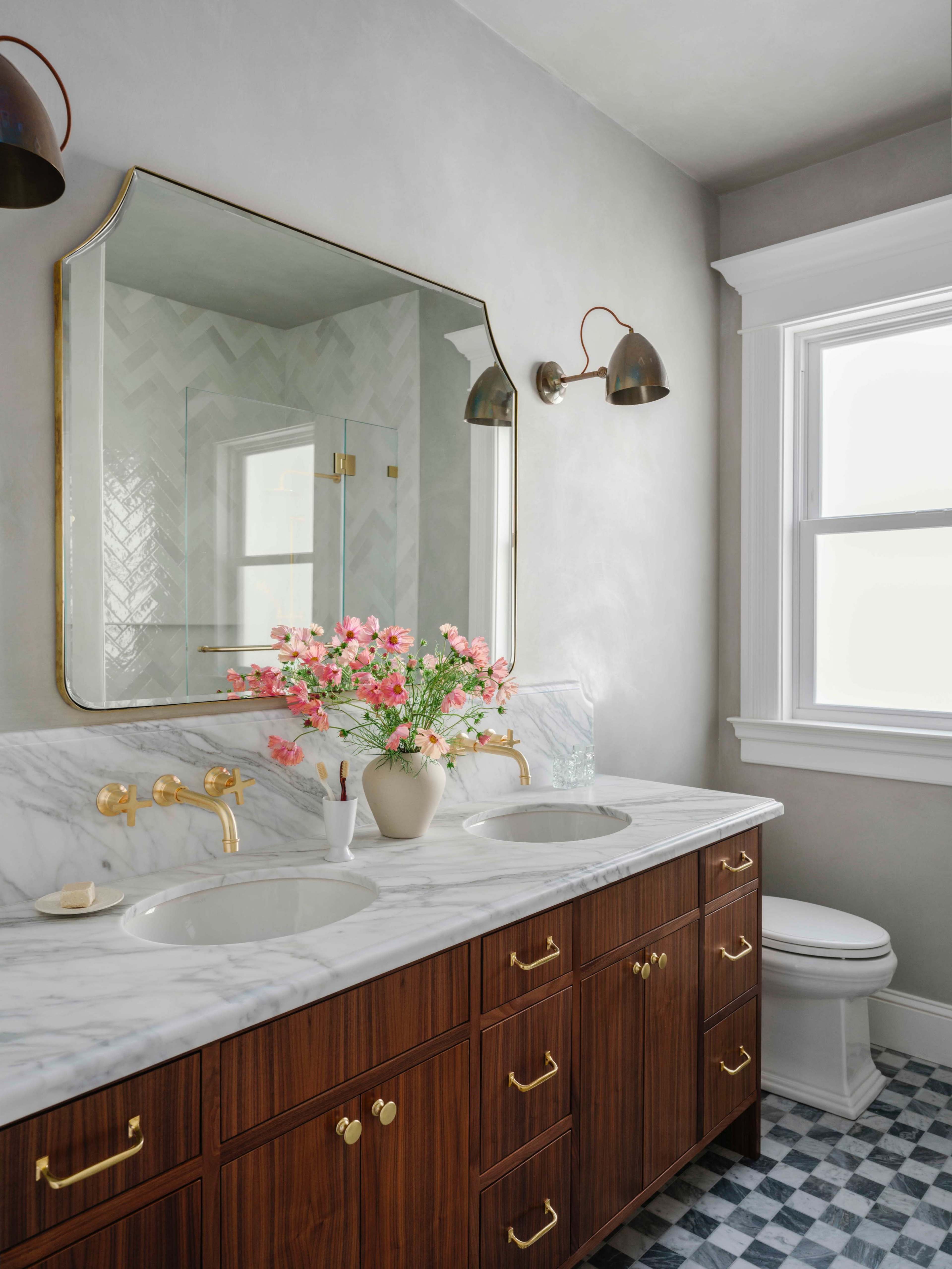 The image shows a modern bathroom with a marble double vanity, a large mirror, wall sconces, and a vase of flowers on the countertop, alongside a toilet and a window with natural light.