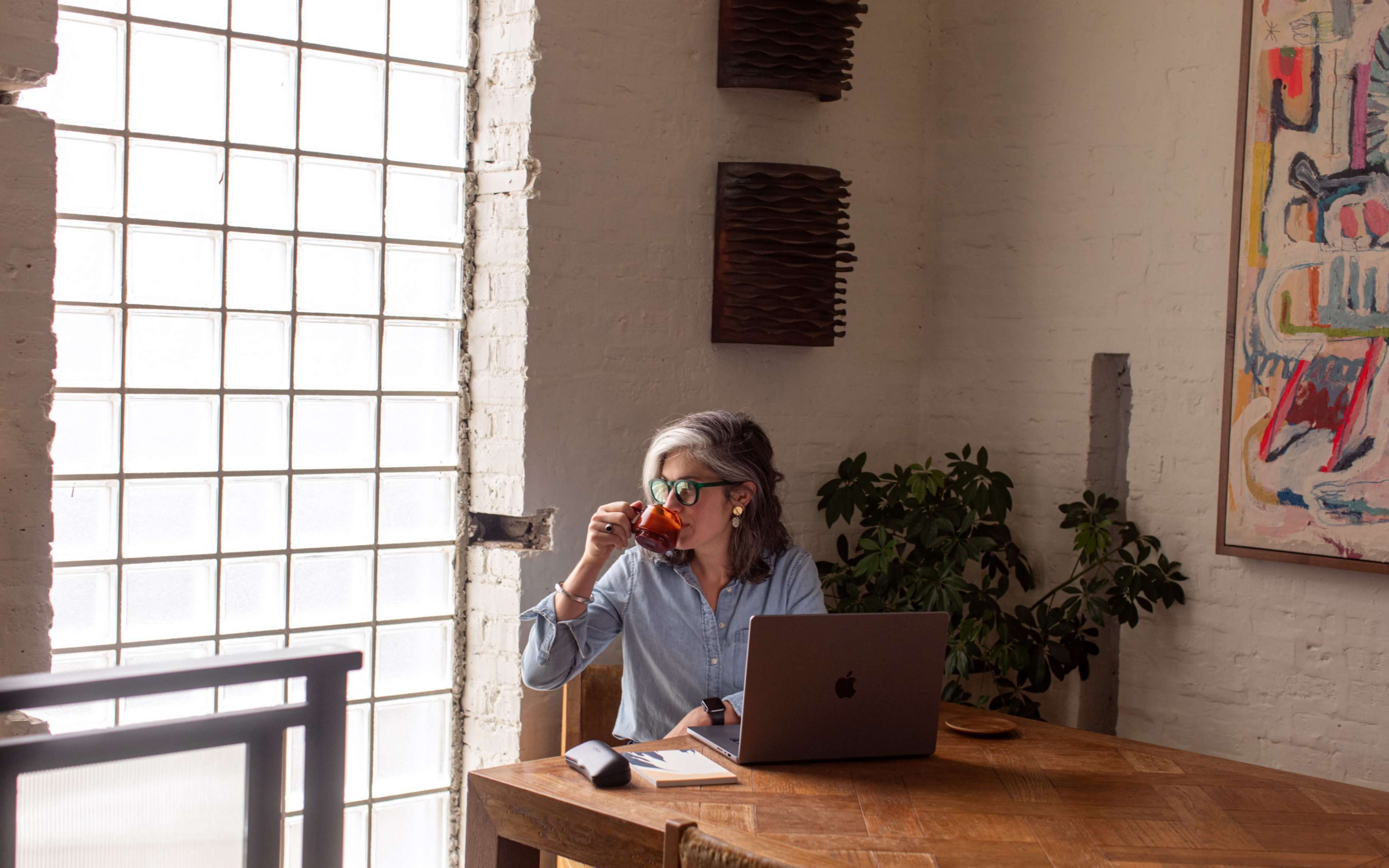 A person sits at a wooden table with a laptop, sipping from a mug near a large window with glass blocks.
