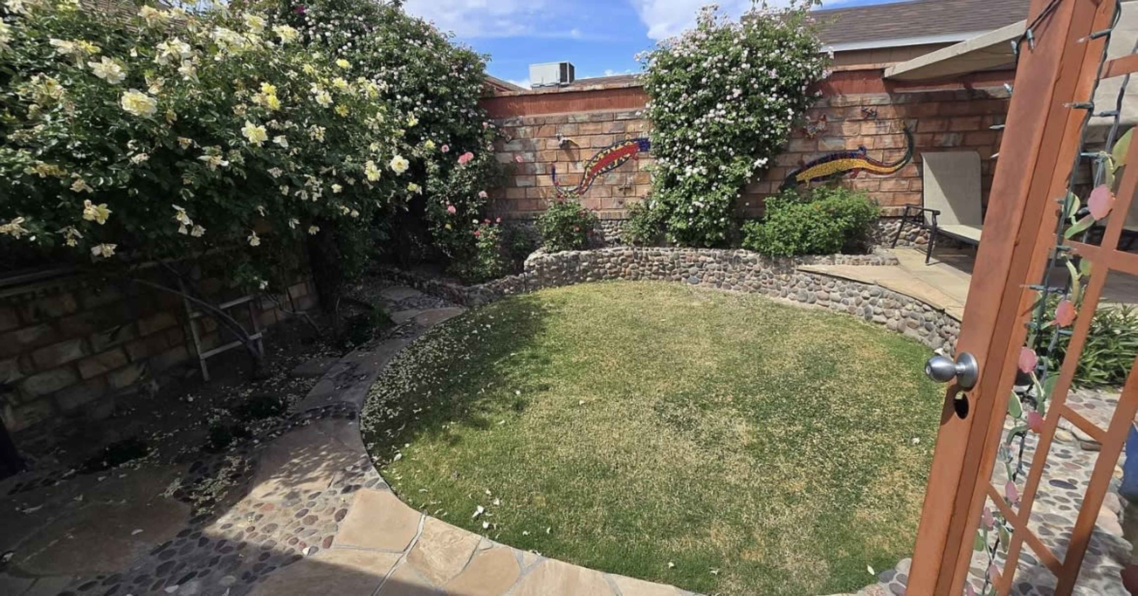 The image shows a circular patch of grass enclosed by stone walls and flowering plants, with a decorative gate in the foreground.