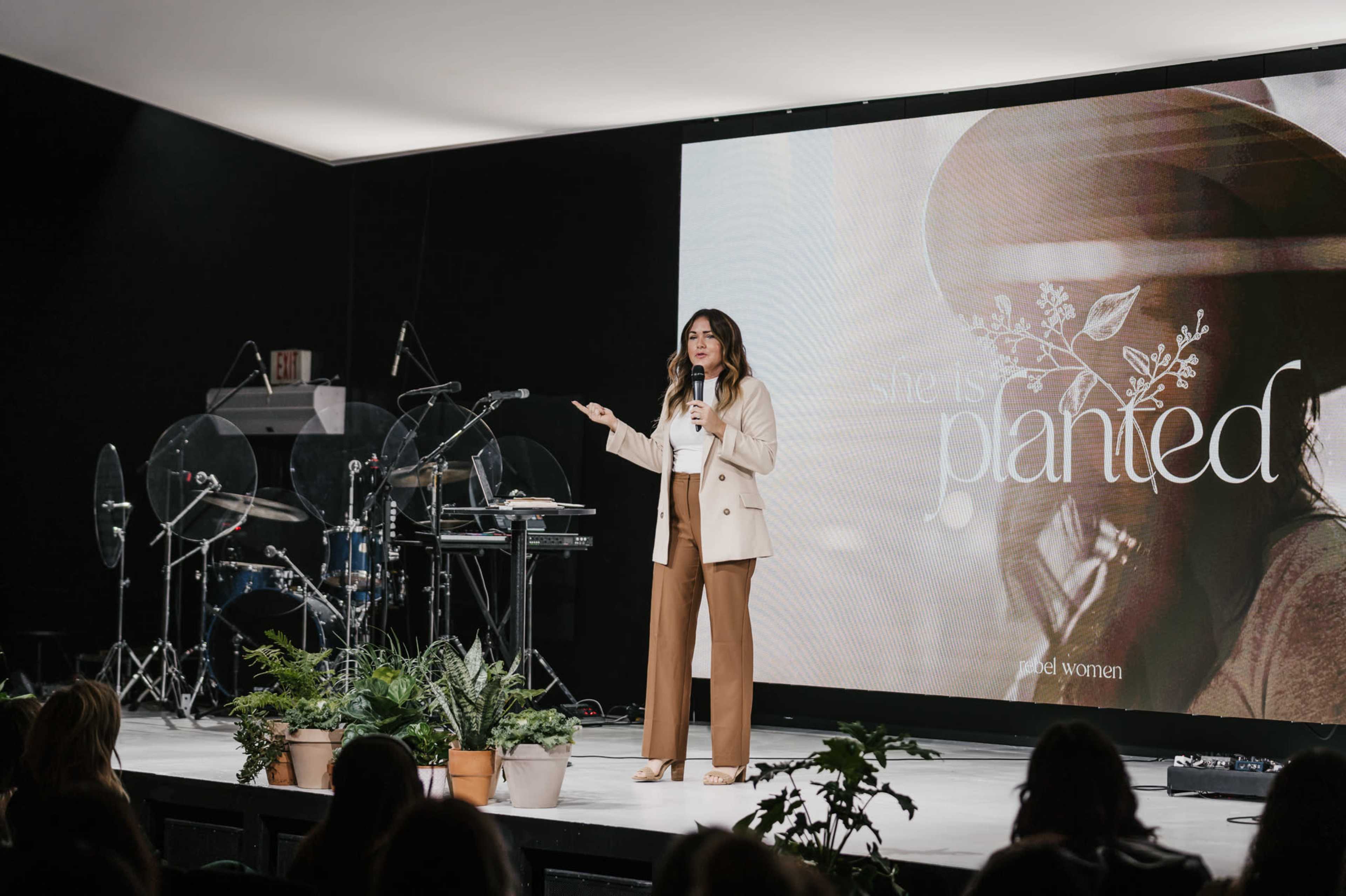 A woman stands on stage, speaking to an audience with a large presentation screen behind her displaying the text "she is planted."