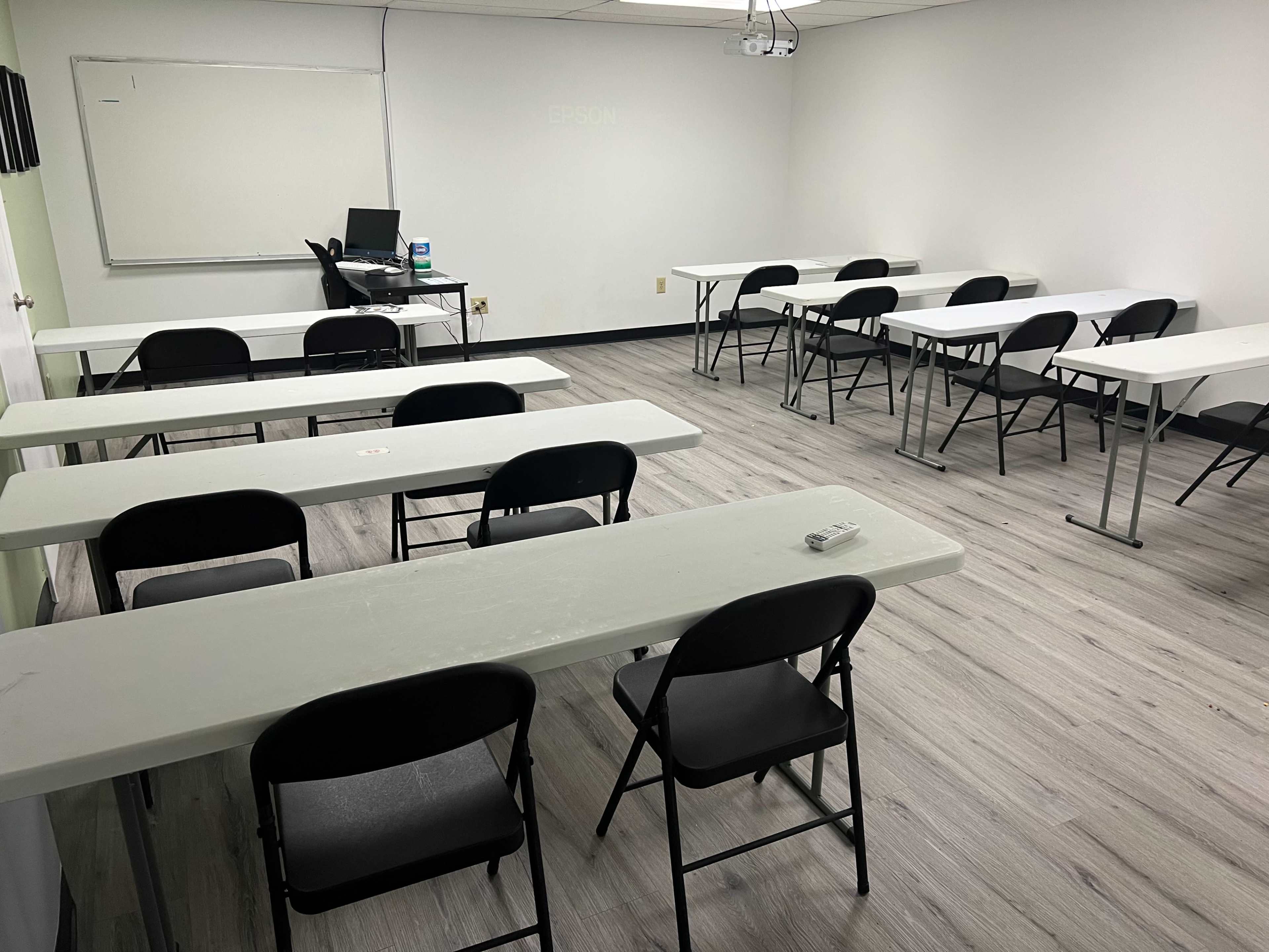 A sparsely furnished classroom with several white tables and black chairs arranged for use.