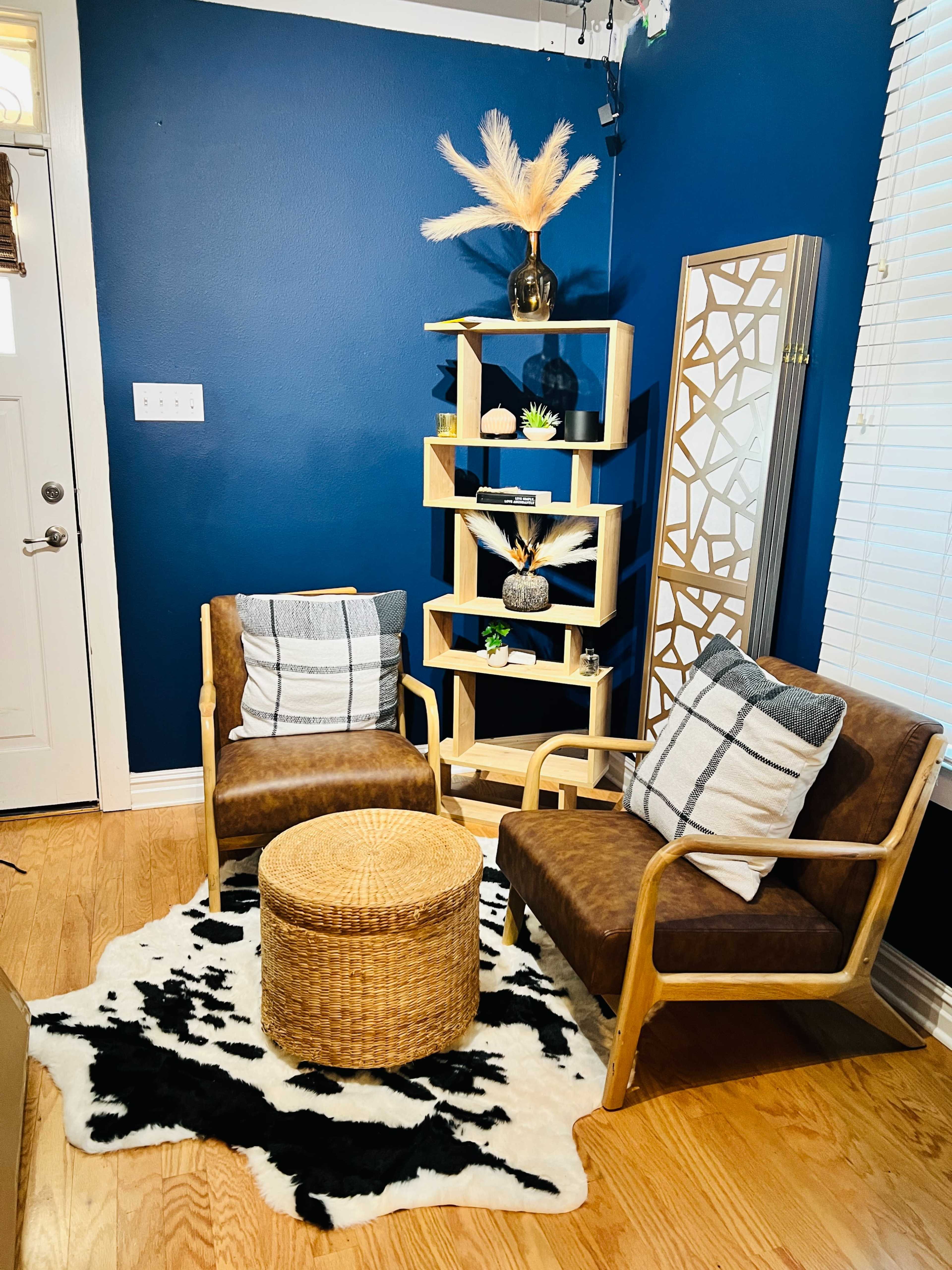 The image shows a cozy living space with two brown chairs, a round woven table, a black and white patterned rug, and a wooden shelf decorated with plants and a vase on a blue wall.