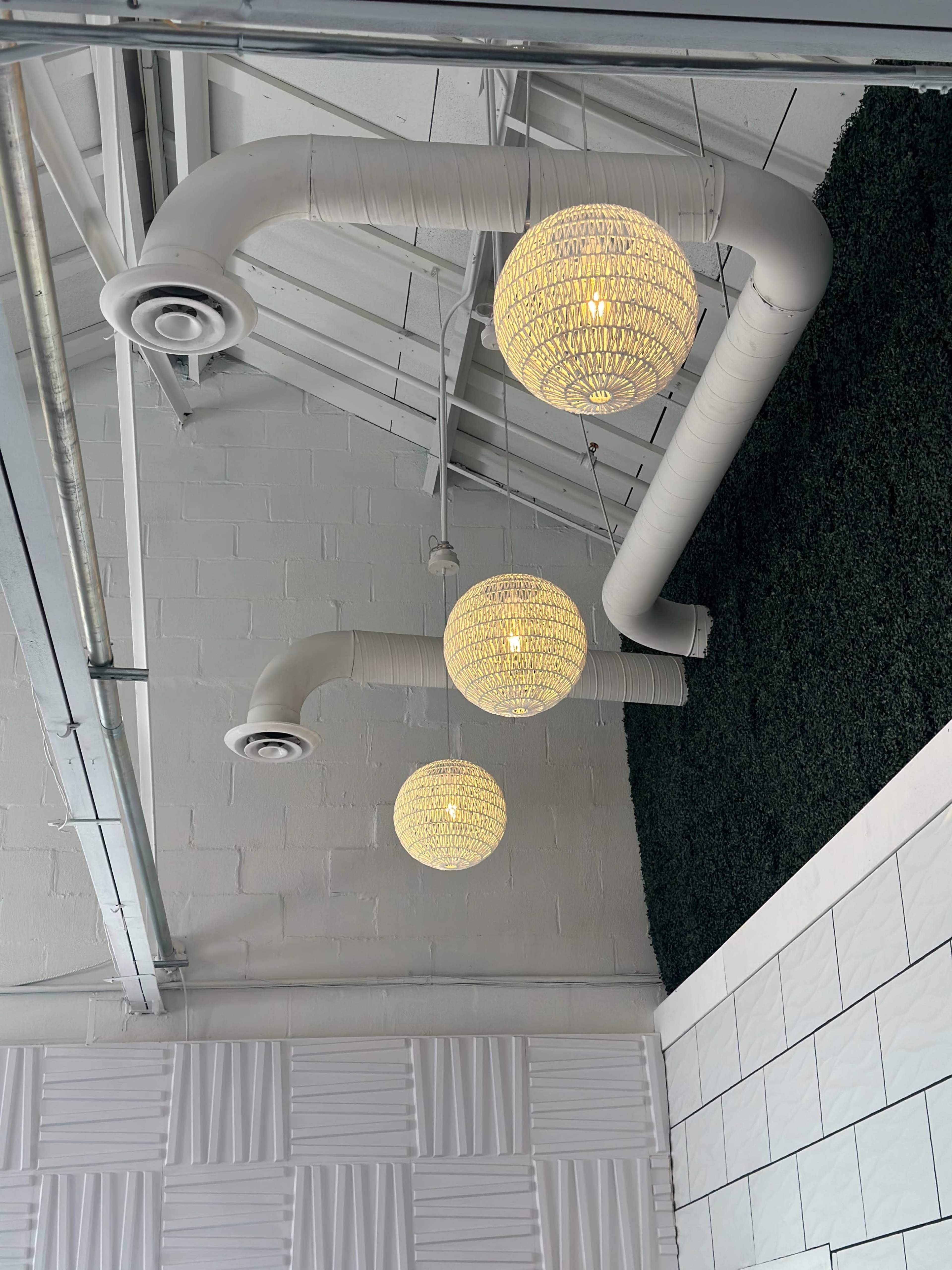 Three pendant lights hang from a ceiling with exposed ductwork and a wall partially covered in artificial grass.