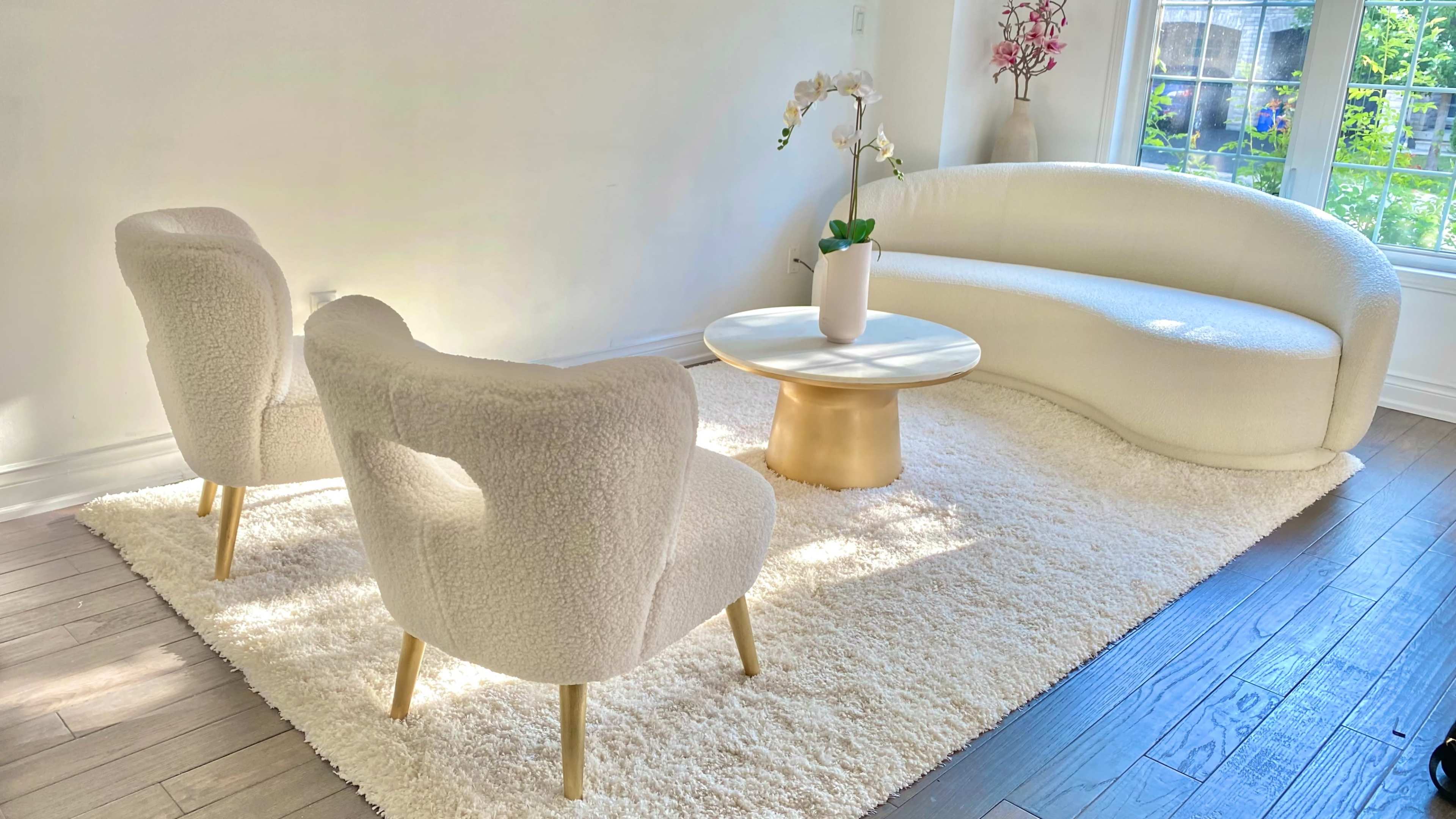 A modern living room features a curved white sofa, two textured chairs, a round table, and an orchid centerpiece on a plush area rug.