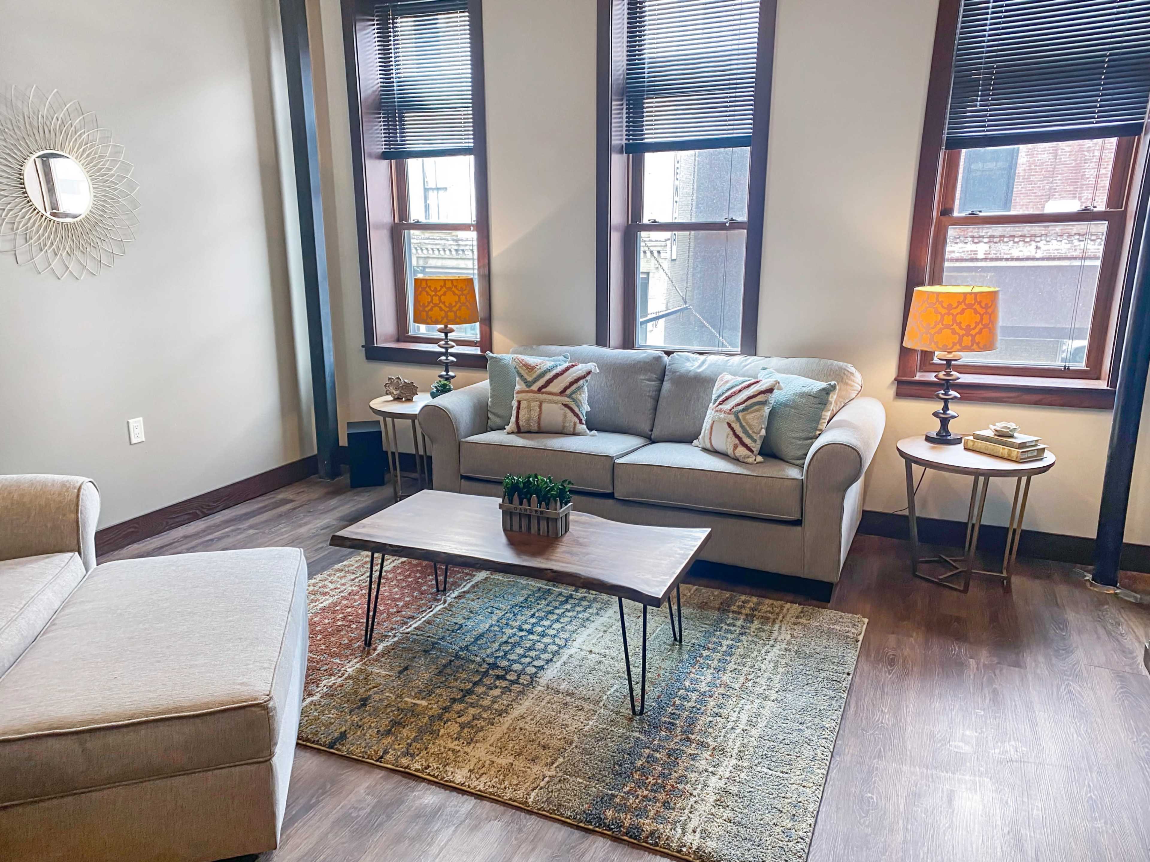 The image shows a modern living room with a gray sofa, a wooden coffee table on a patterned rug, and two lamps on side tables near large windows.