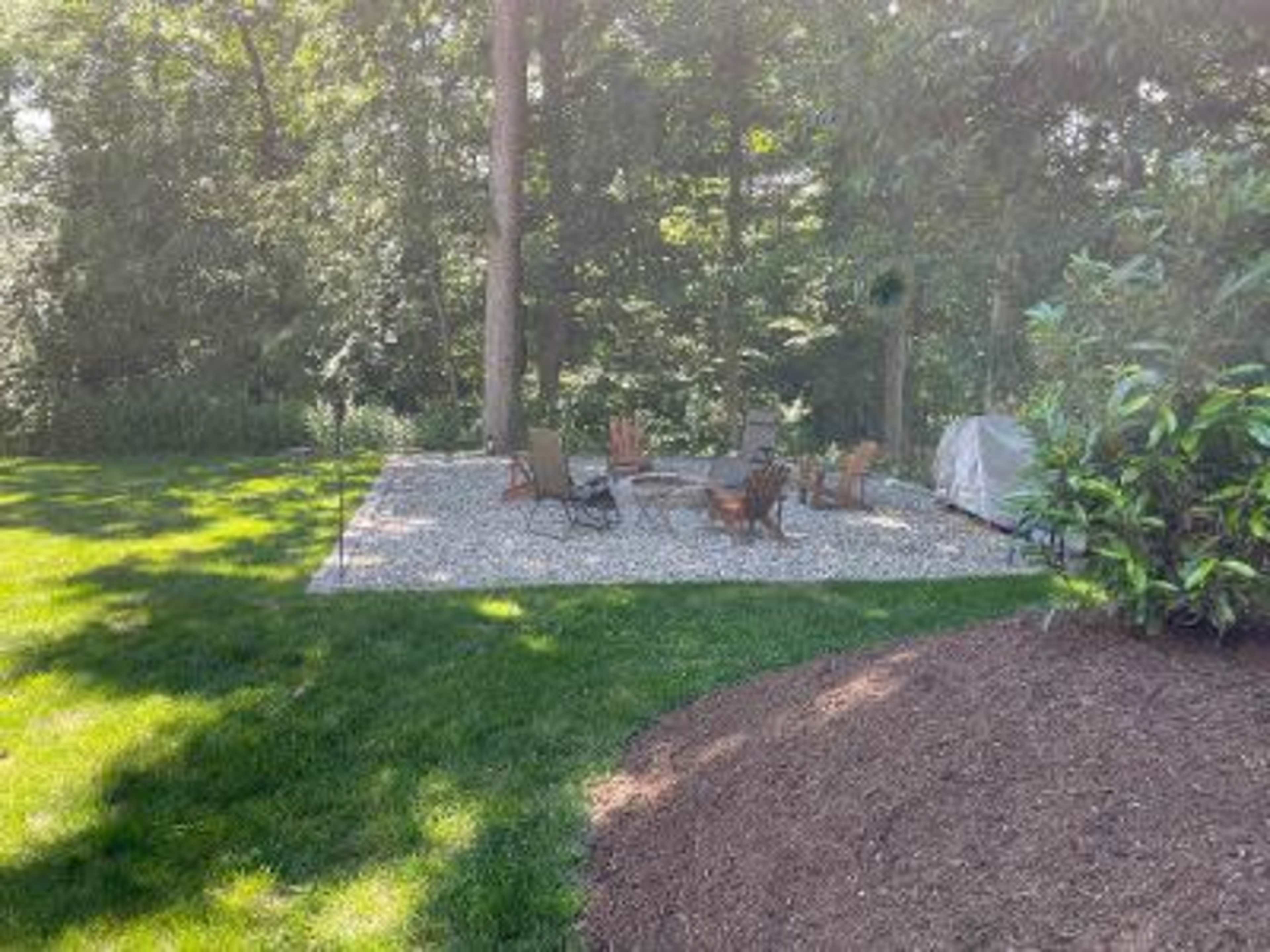 A gravel patio surrounded by trees features several lawn chairs arranged around a fire pit.