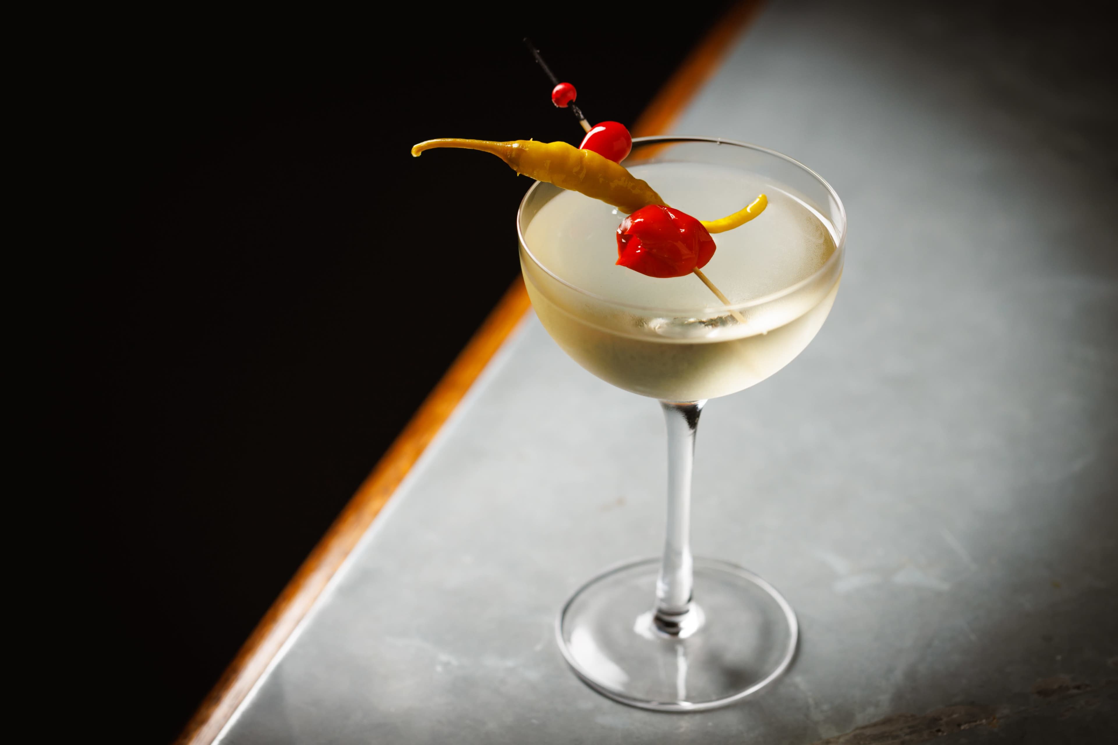 A cocktail with a clear liquid is served in a elegant glass and garnished with a yellow chili pepper and red cherries on a toothpick.