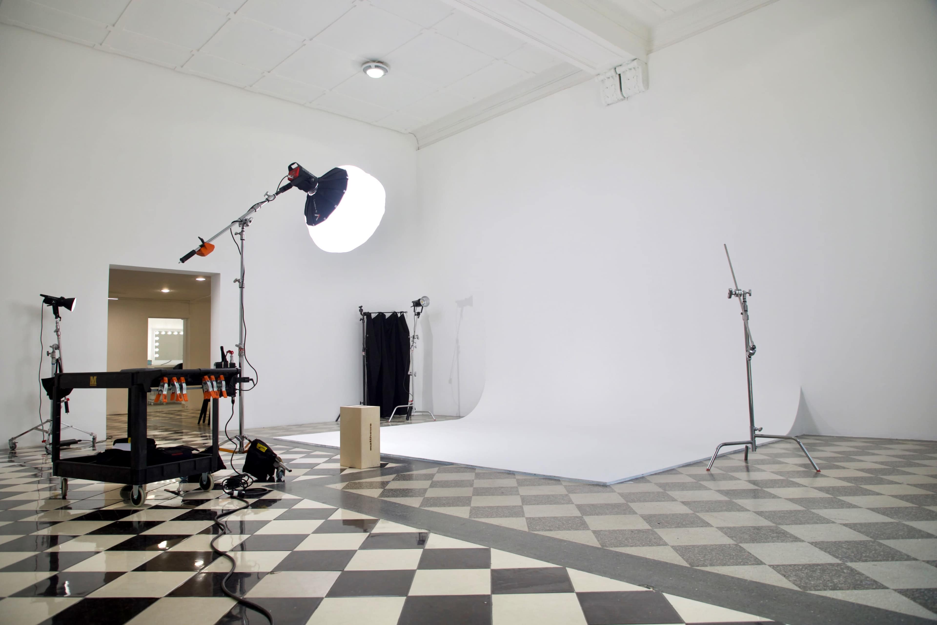 The image shows a photography studio with a white seamless backdrop, lighting equipment, and a checkered floor.