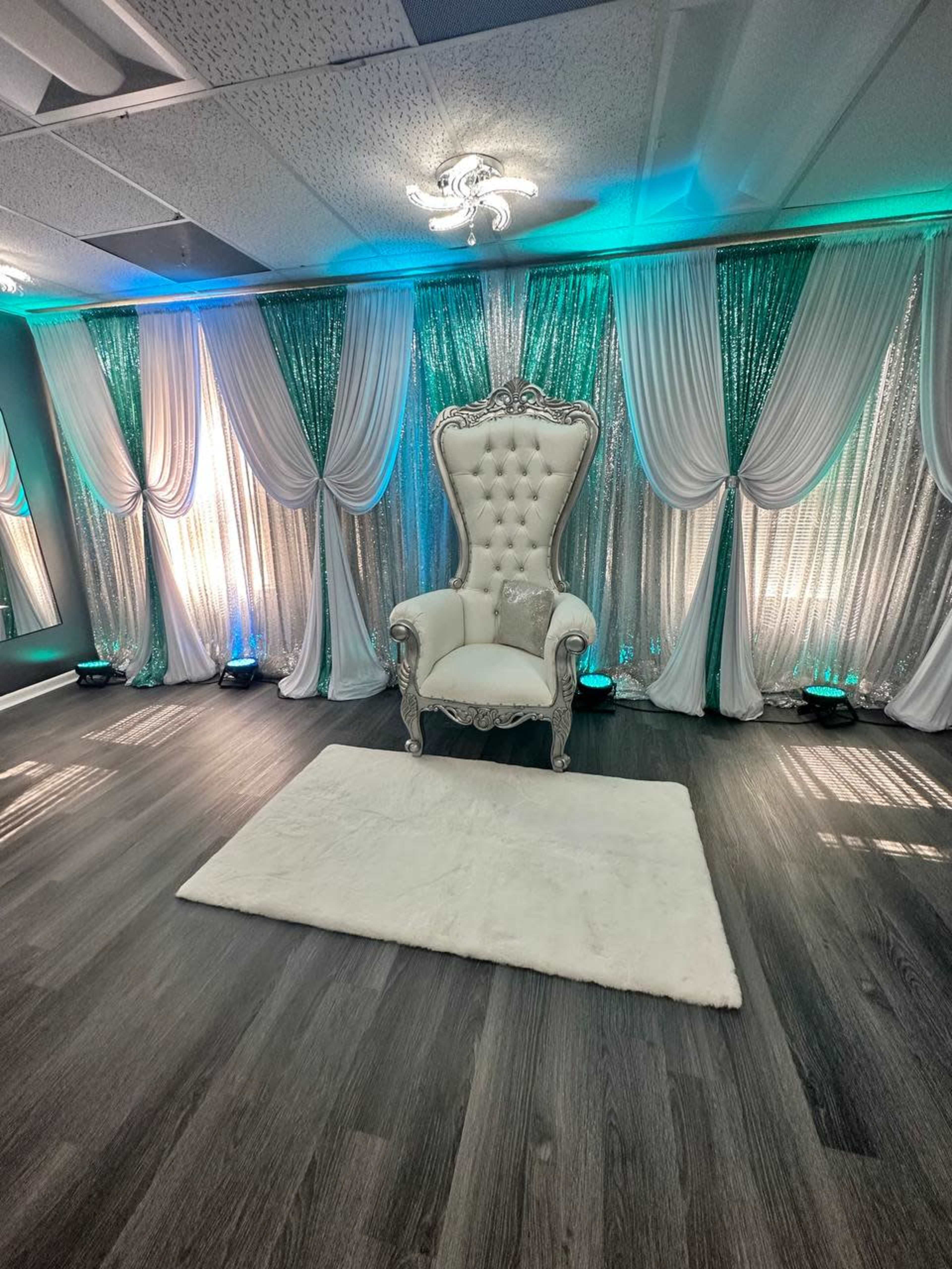 A decorated indoor space featuring a large, ornate silver chair placed on a fluffy white rug, surrounded by sequined backdrops and soft lighting.