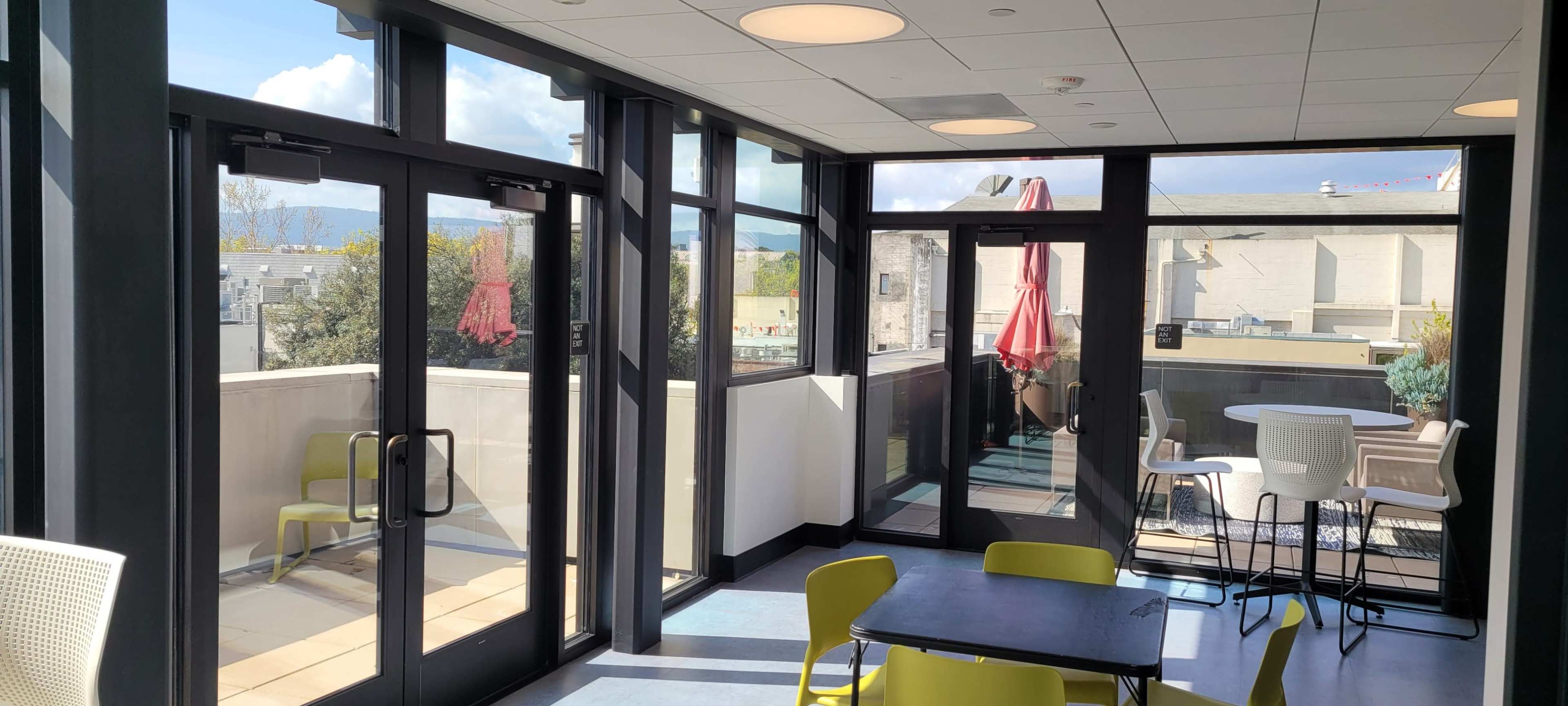 The image shows a modern indoor seating area with large windows, a table with chairs, and a door leading to an outdoor balcony featuring an umbrella.