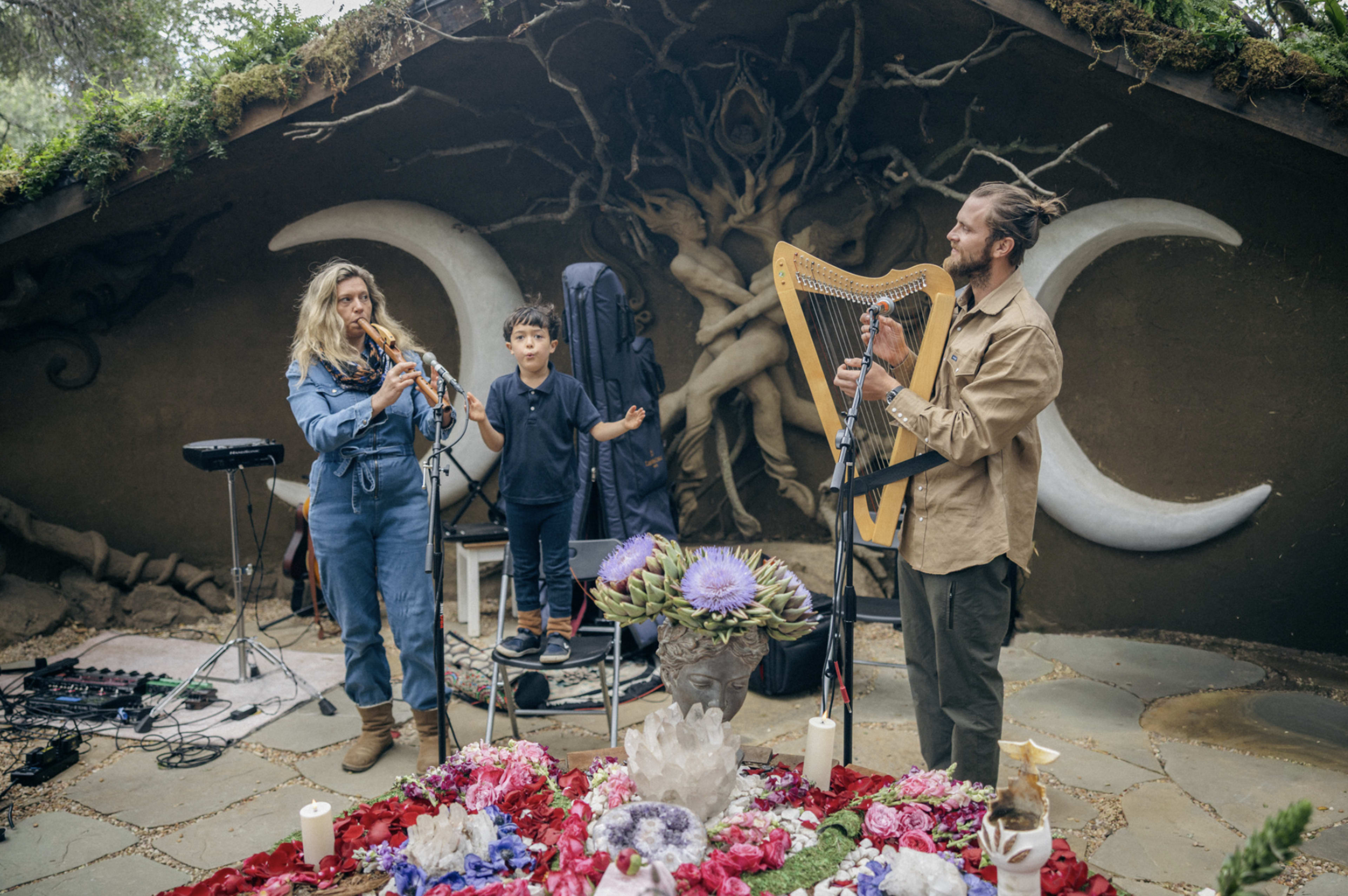 A woman plays a flute, a child stands between her and a man playing a harp, all performing in front of a decorative backdrop featuring crescent moon motifs and floral arrangements on the ground.