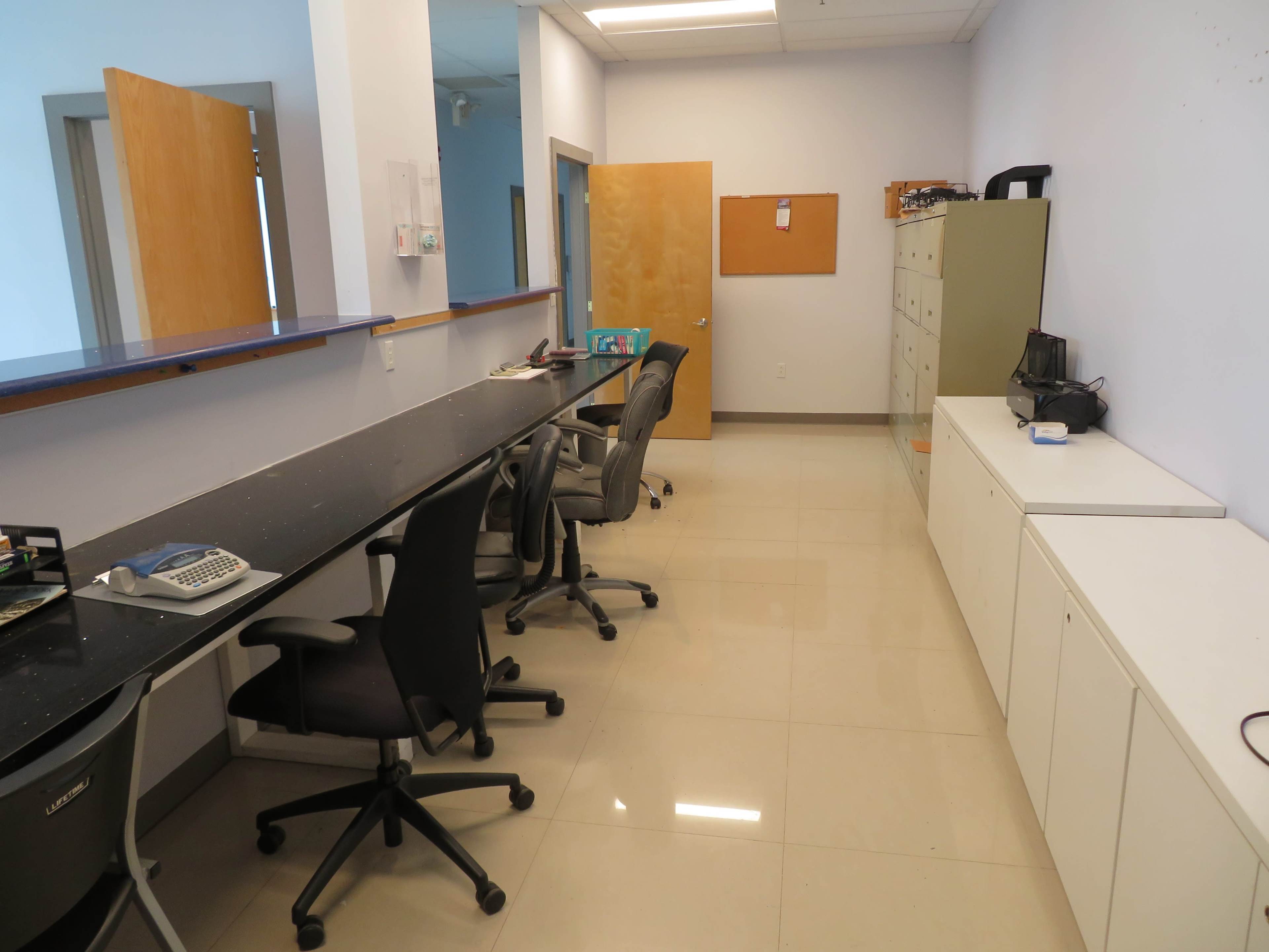The image shows an office space with a long black desk, several office chairs, and storage cabinets lined against the wall.