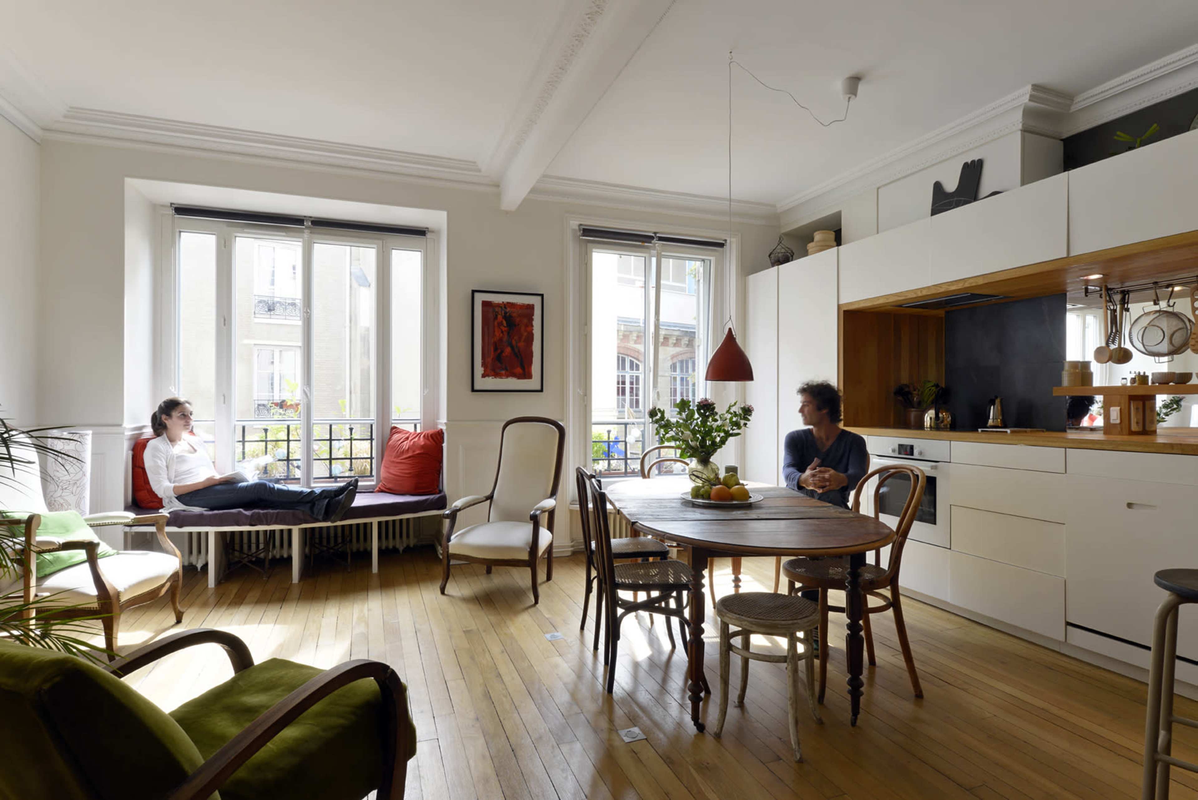 A bright, modern kitchen and dining area features a round wooden table, a bench by the window, and two people seated in the space.