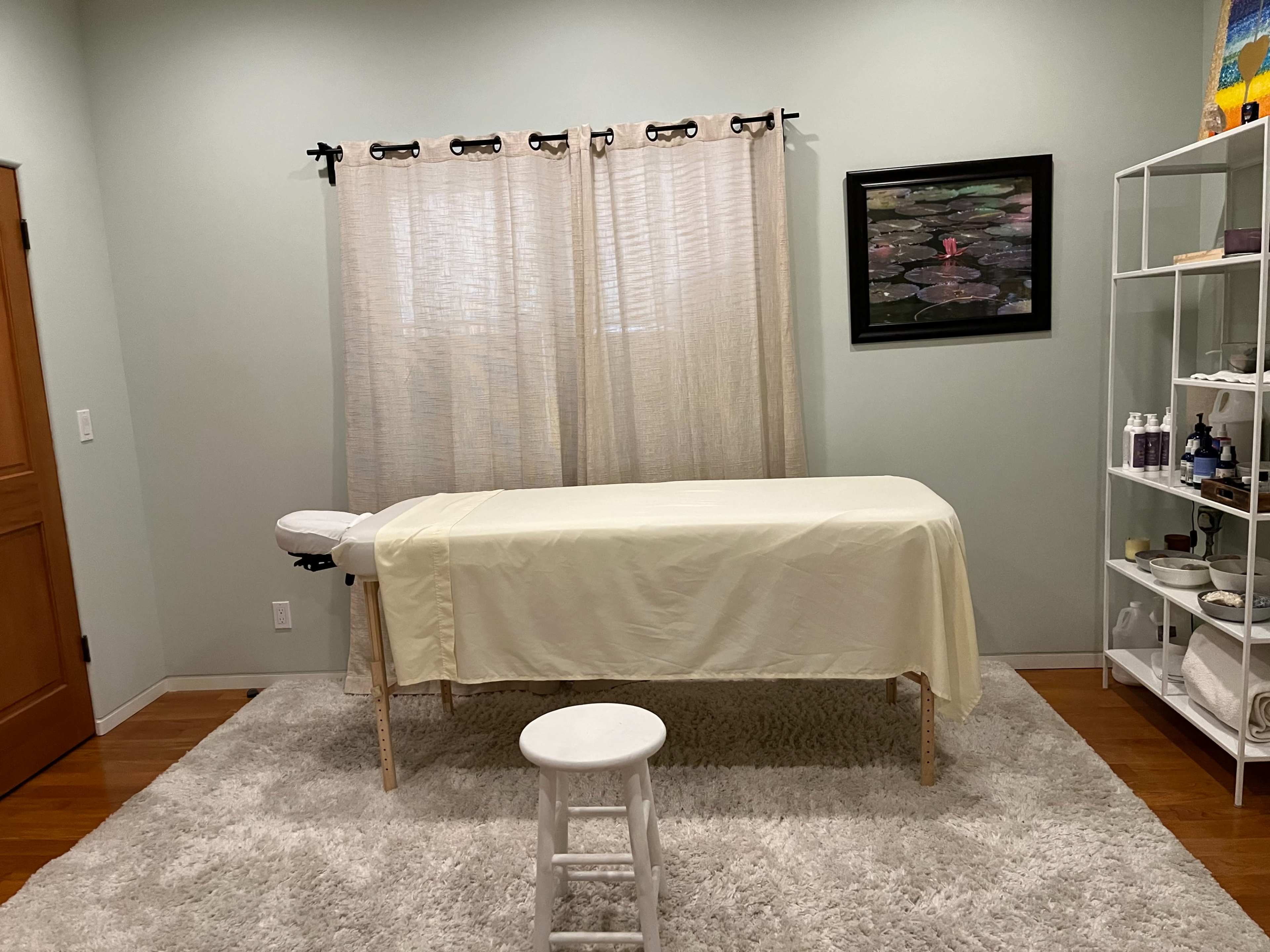 A massage therapy room features a massage table covered with a cloth, a white stool, and curtains allowing light to enter, along with a few shelves containing various supplies.