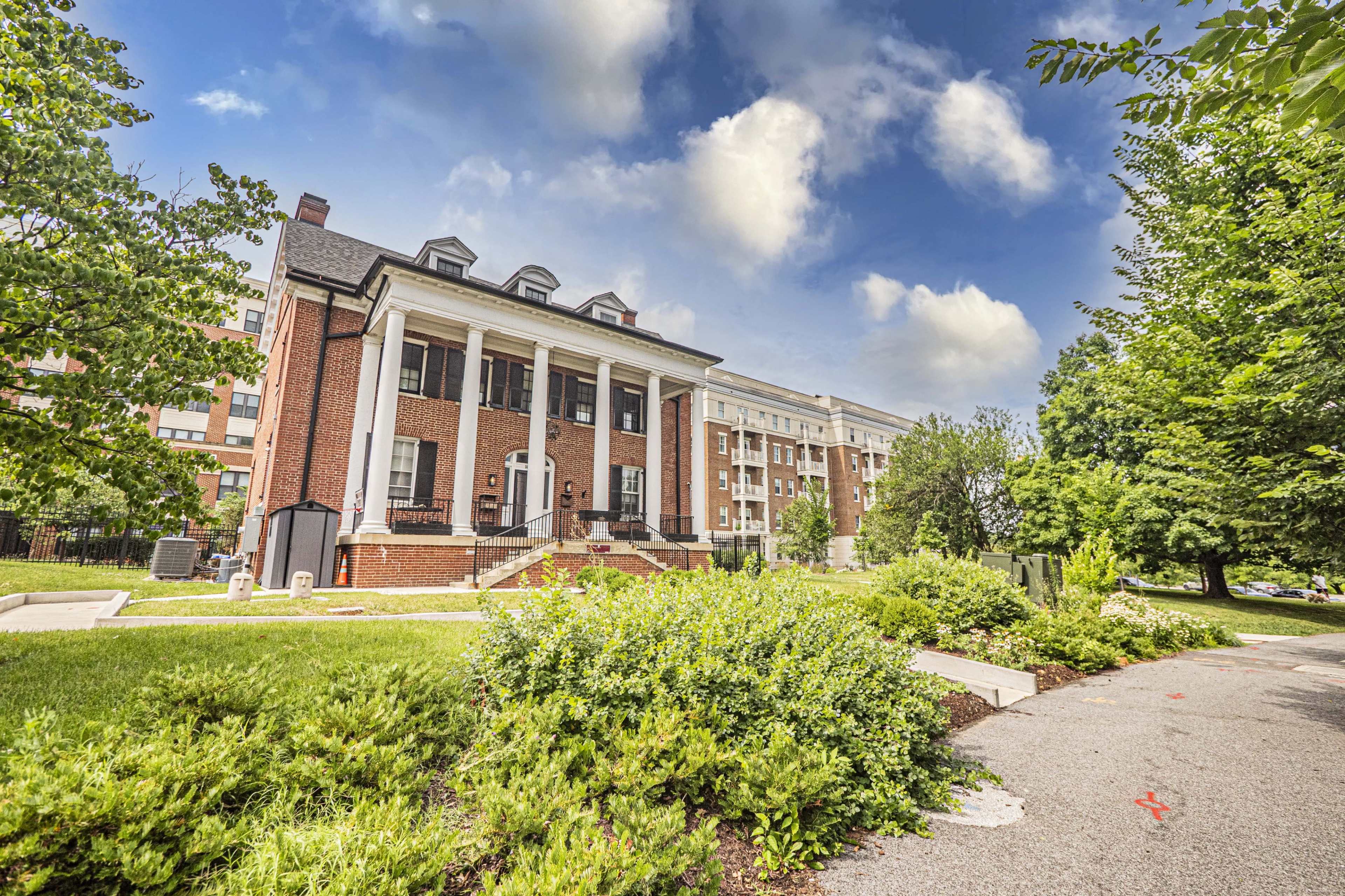The image shows a large brick building with a white columned entrance surrounded by greenery and a paved path.