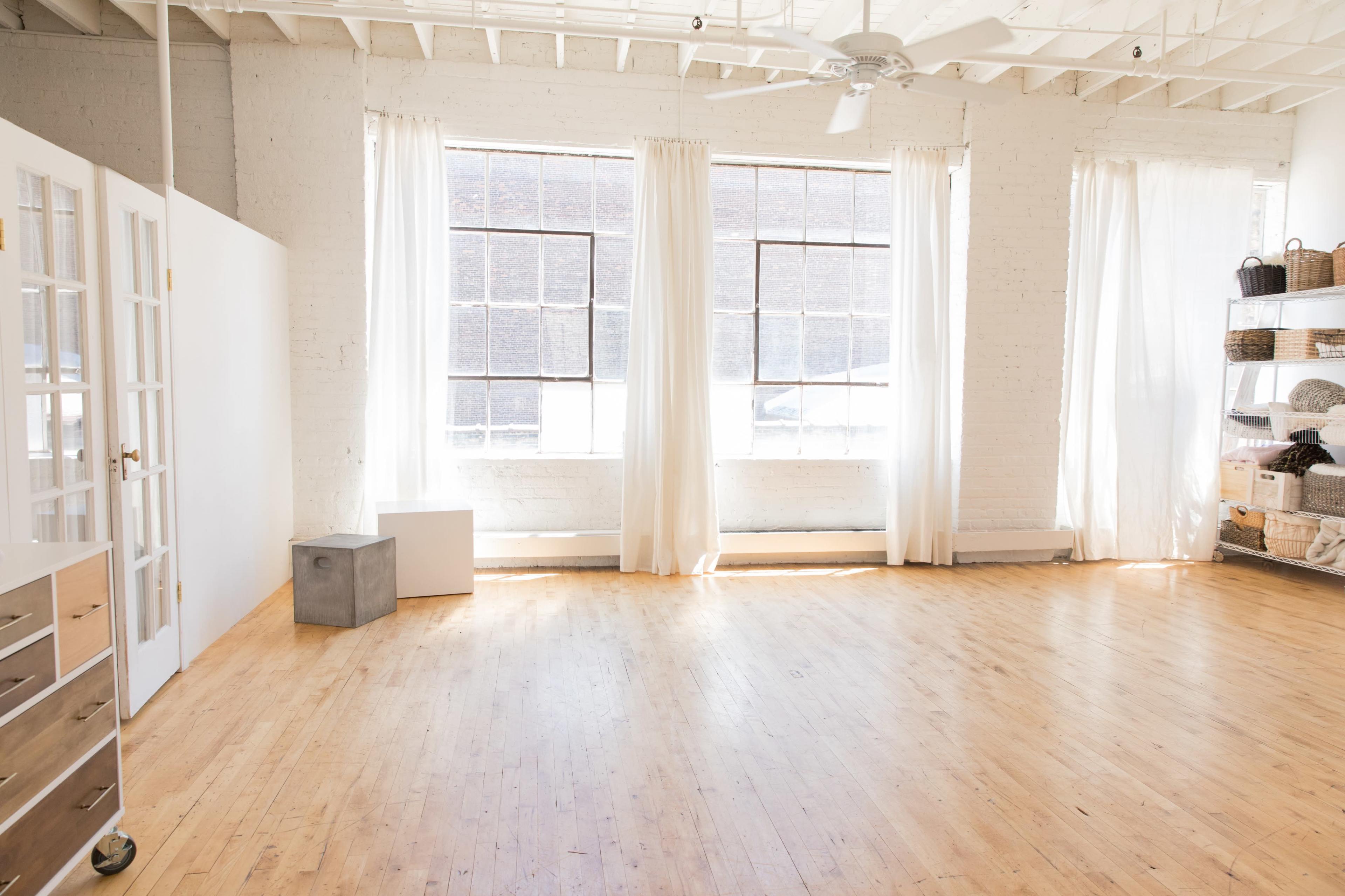 A spacious room with wooden floors, large windows dressed with white curtains, and minimal furnishings including a white cabinet and metal storage unit.